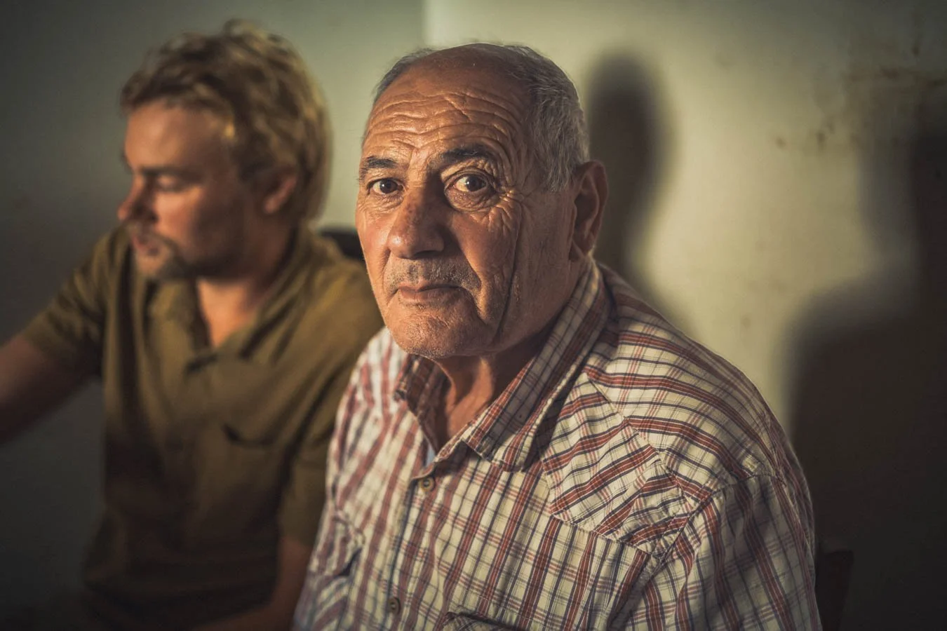 Twee oudere mannen zitten aan een tafel in een kamer, met één man die direct in de camera kijkt terwijl de andere zich afwendt.