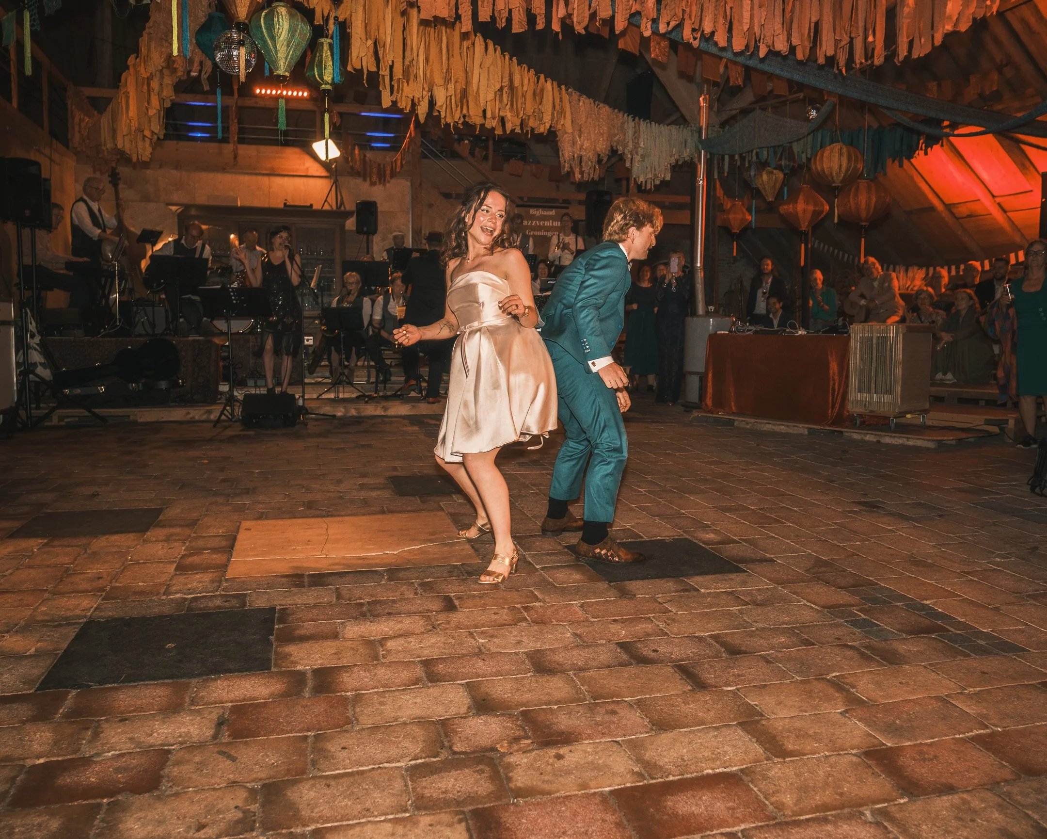 Mannen en vrouw dansen samen op de dansvloer tijdens een feestje, met een band of orkest op de achtergrond in een versierde locatie.