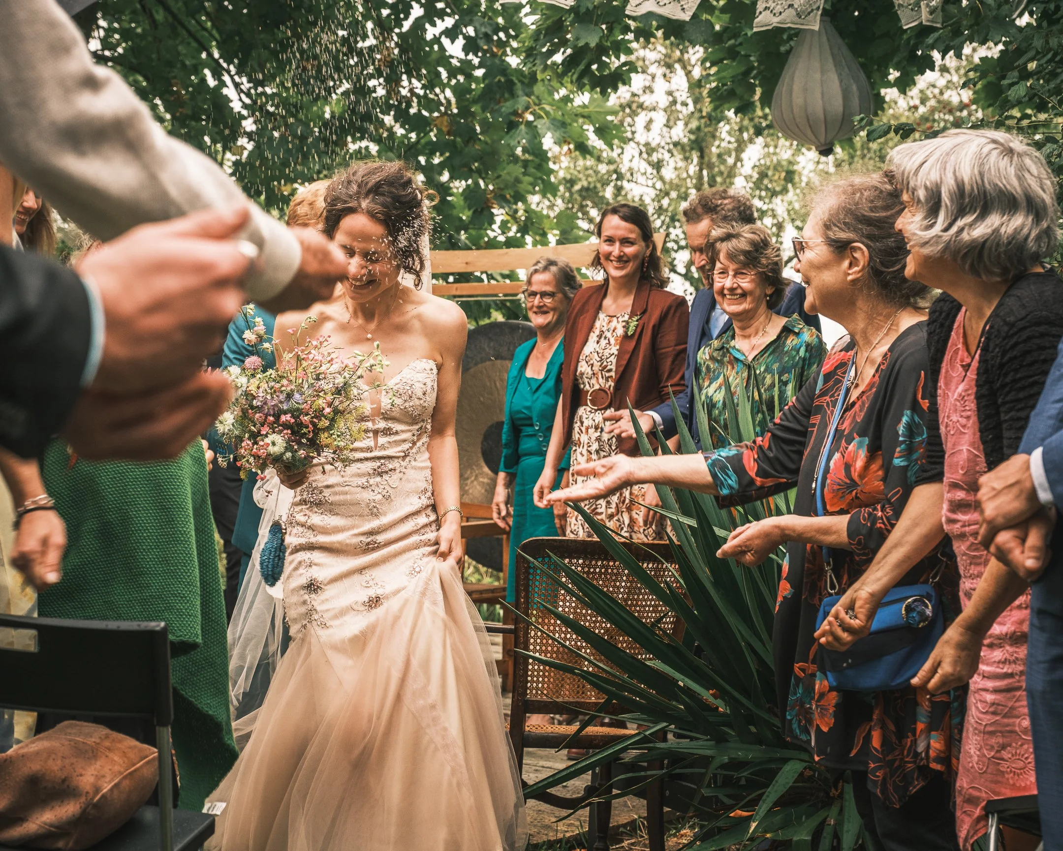 Bruidsmeisje met bloemboeket wordt besproeid met champagne terwijl ze omringd is door lachende gasten op een tuinbruiloft.