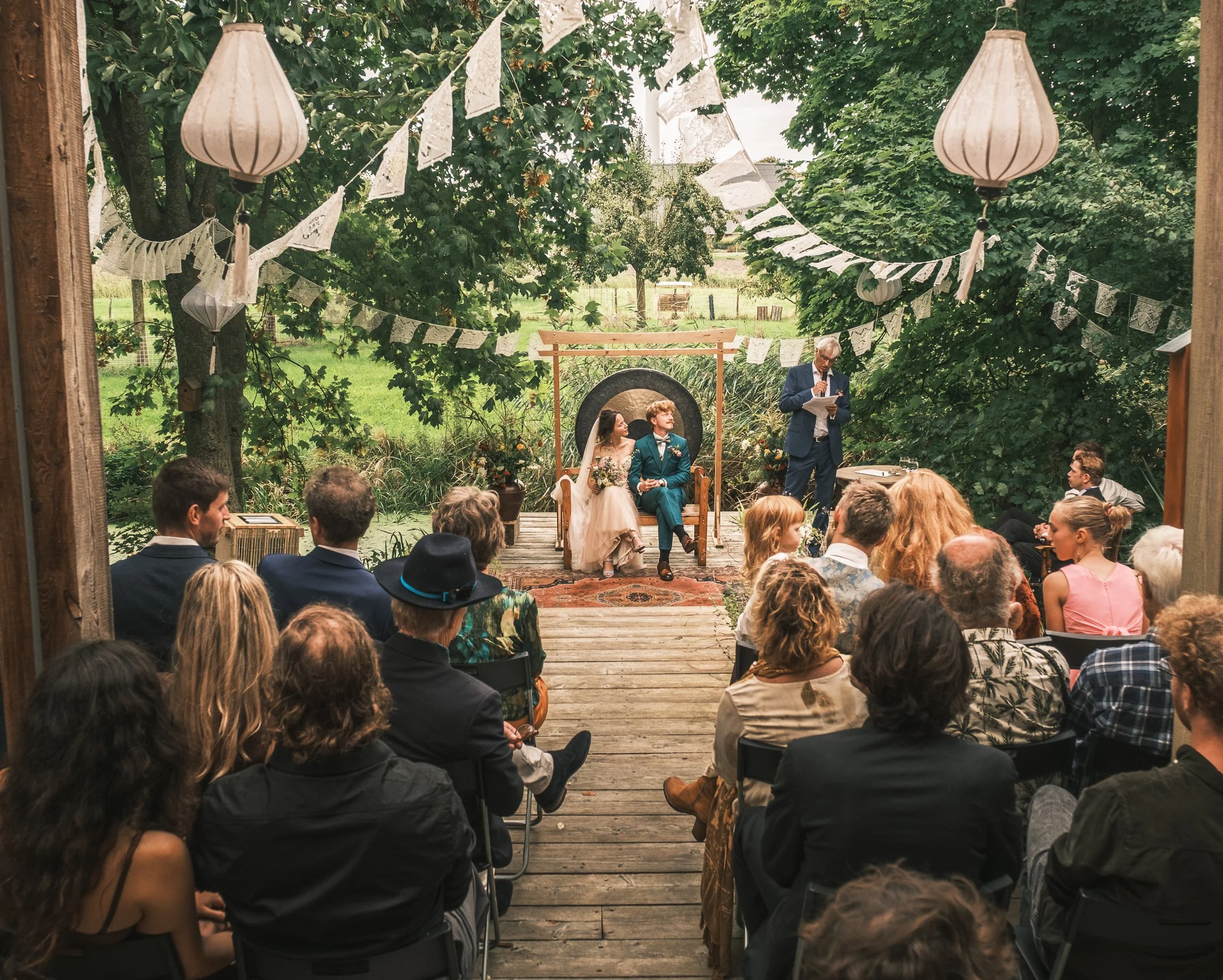 Een bruiloftceremonie onder een groene boog, met gasten die kijken naar het bruidspaar dat op een verhoging zit, terwijl iemand voordraagt voor de trouwgeloften, in een natuurlijke buitenomgeving versierd met papieren lampen en vlaggen.