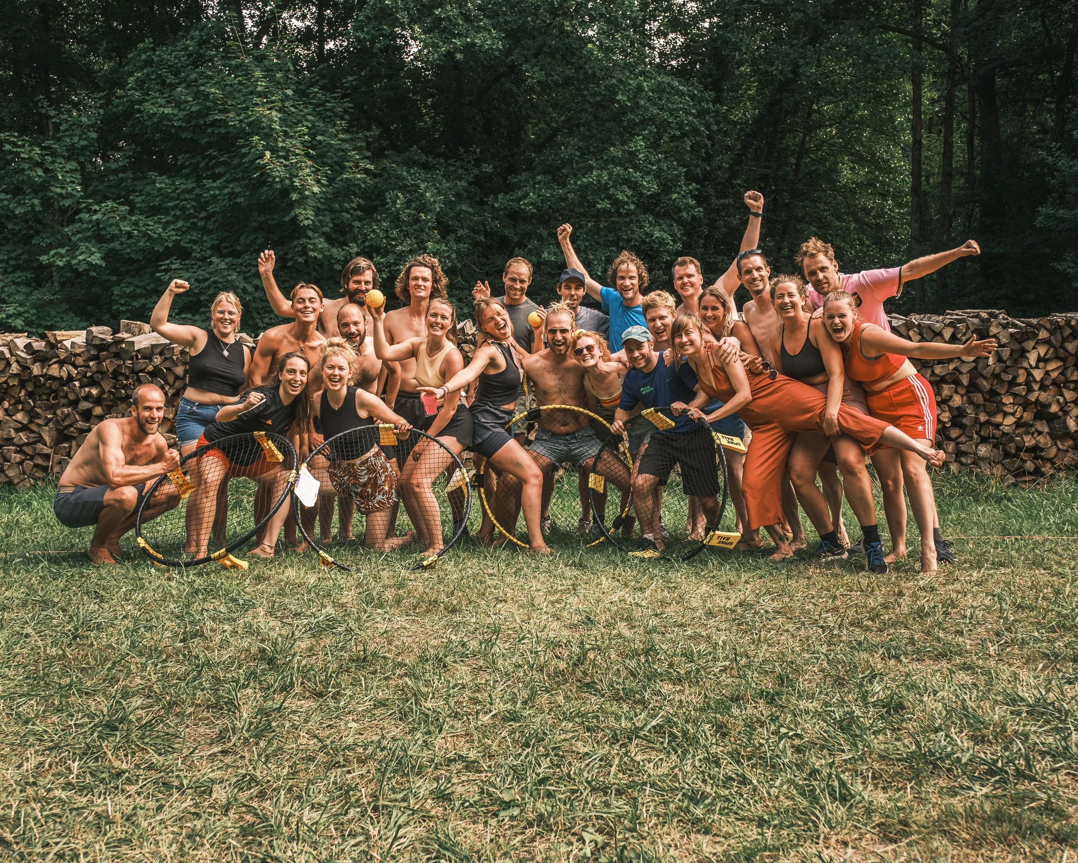 Een grote groep mensen die buiten in een bosrijke omgeving plezier heeft, met enkele vrouwen en mannen die yoga hoepels vasthouden en lachend poseert.