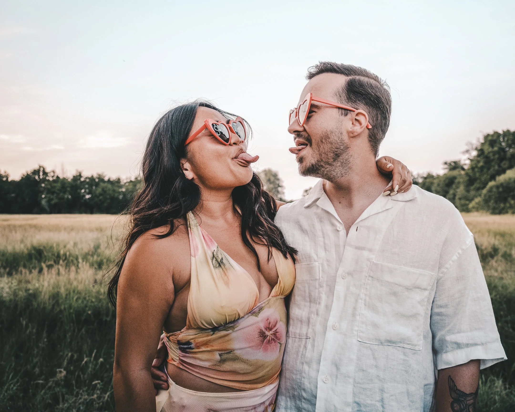 Een man en vrouw in een open veld dragen hartvormige zonnebrillen, maken een speelse houding en kijken naar elkaar.