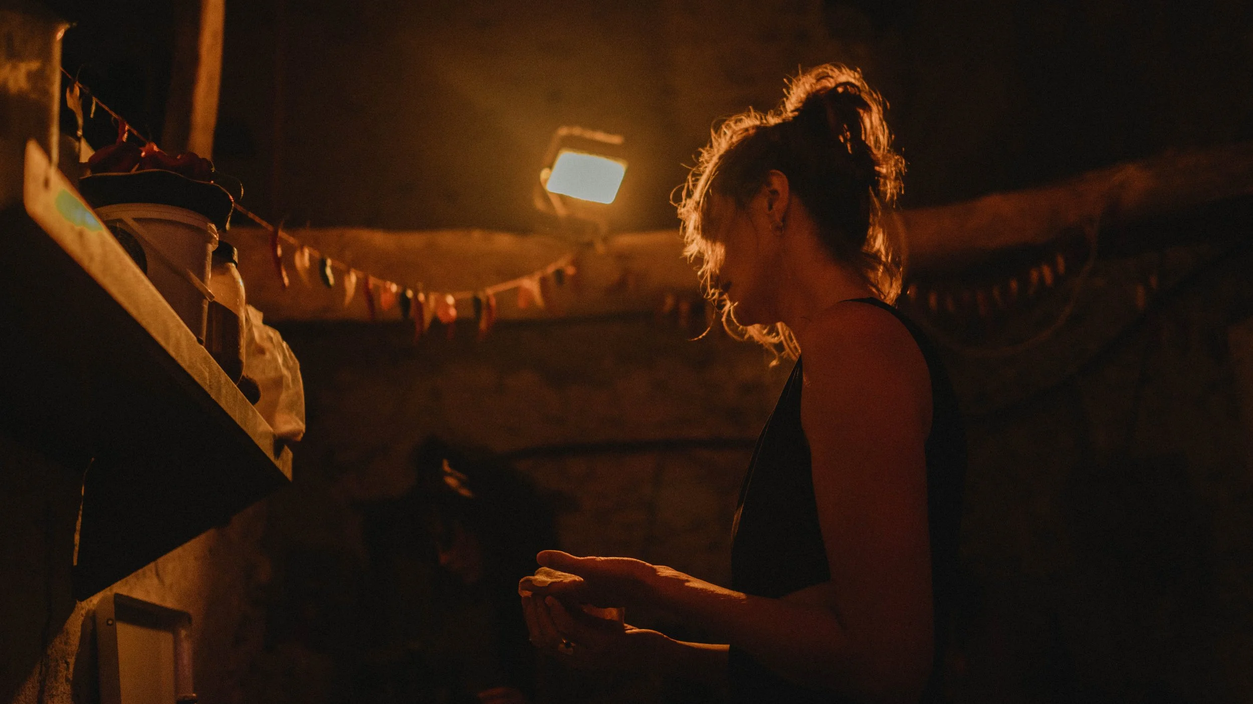 Een vrouw met krulla haar in een donkere kamer, verlicht door een warm licht, kijkt naar haar handen.