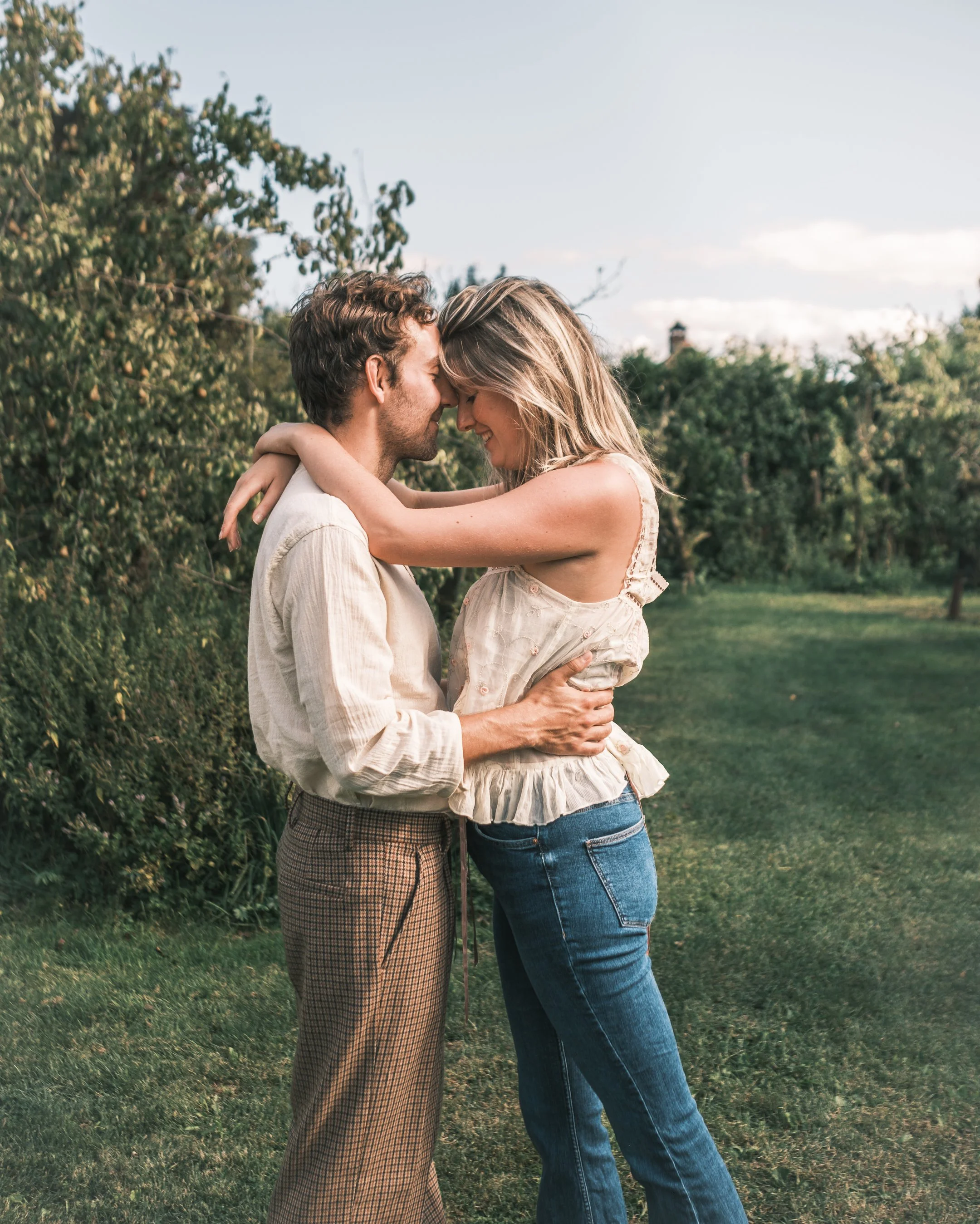Een man en vrouw omhelzen elkaar in een groene tuin, hun voorhoofden raken elkaar en ze lachen, met bomen en een blauwe lucht op de achtergrond.