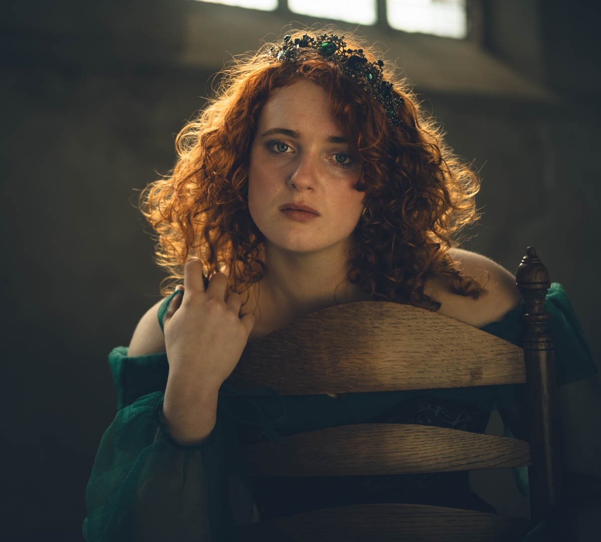 Een jonge vrouw met rood, gekruld haar draagt een tiara en zit achter een houten stoel, met een contemplatieve uitdrukking in een donkere kamer met natuurlijk licht.