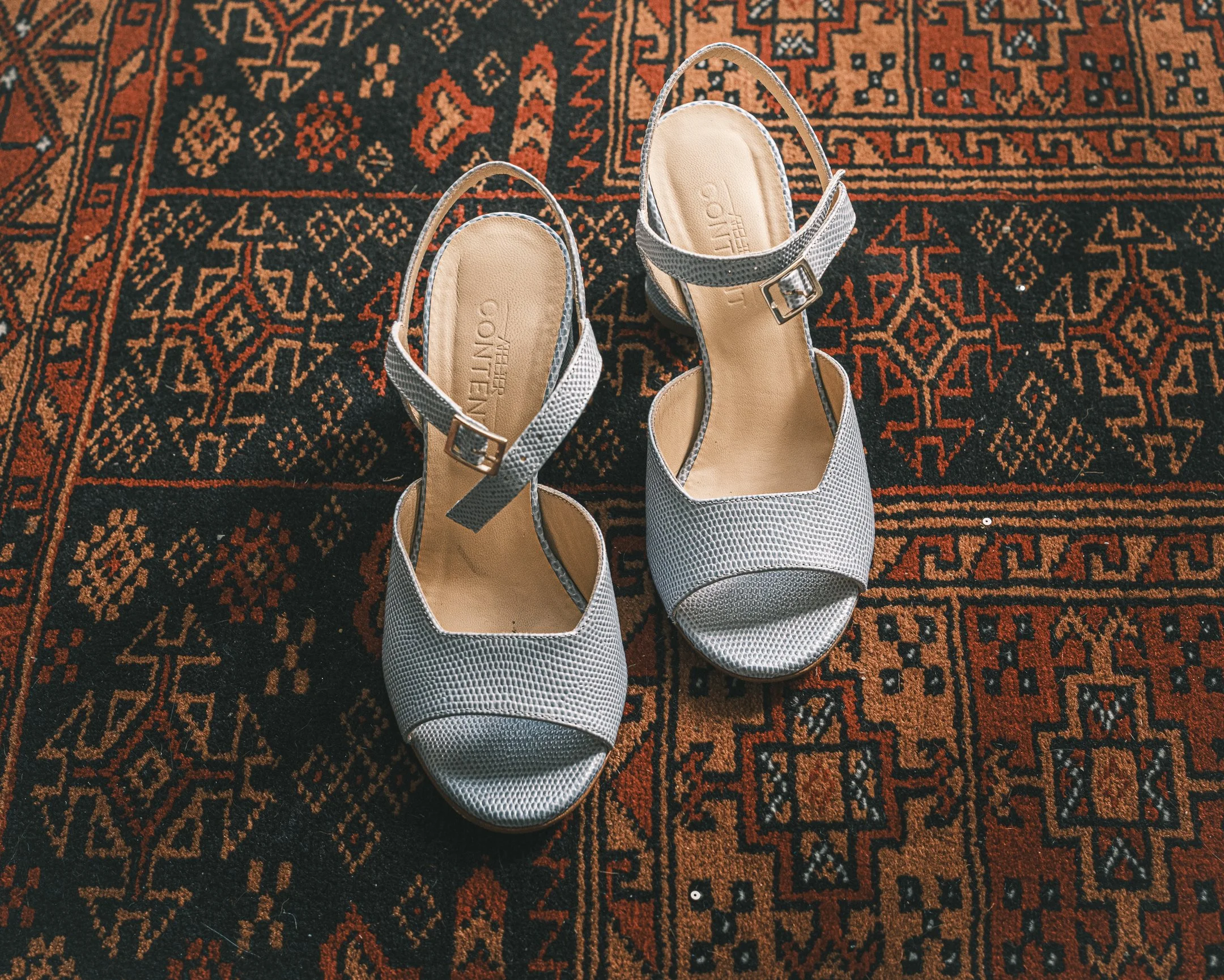 Witte open-toe heeled sandalen met een gespsluiting, geplaatst op een rood en zwart traditioneel tapijt.