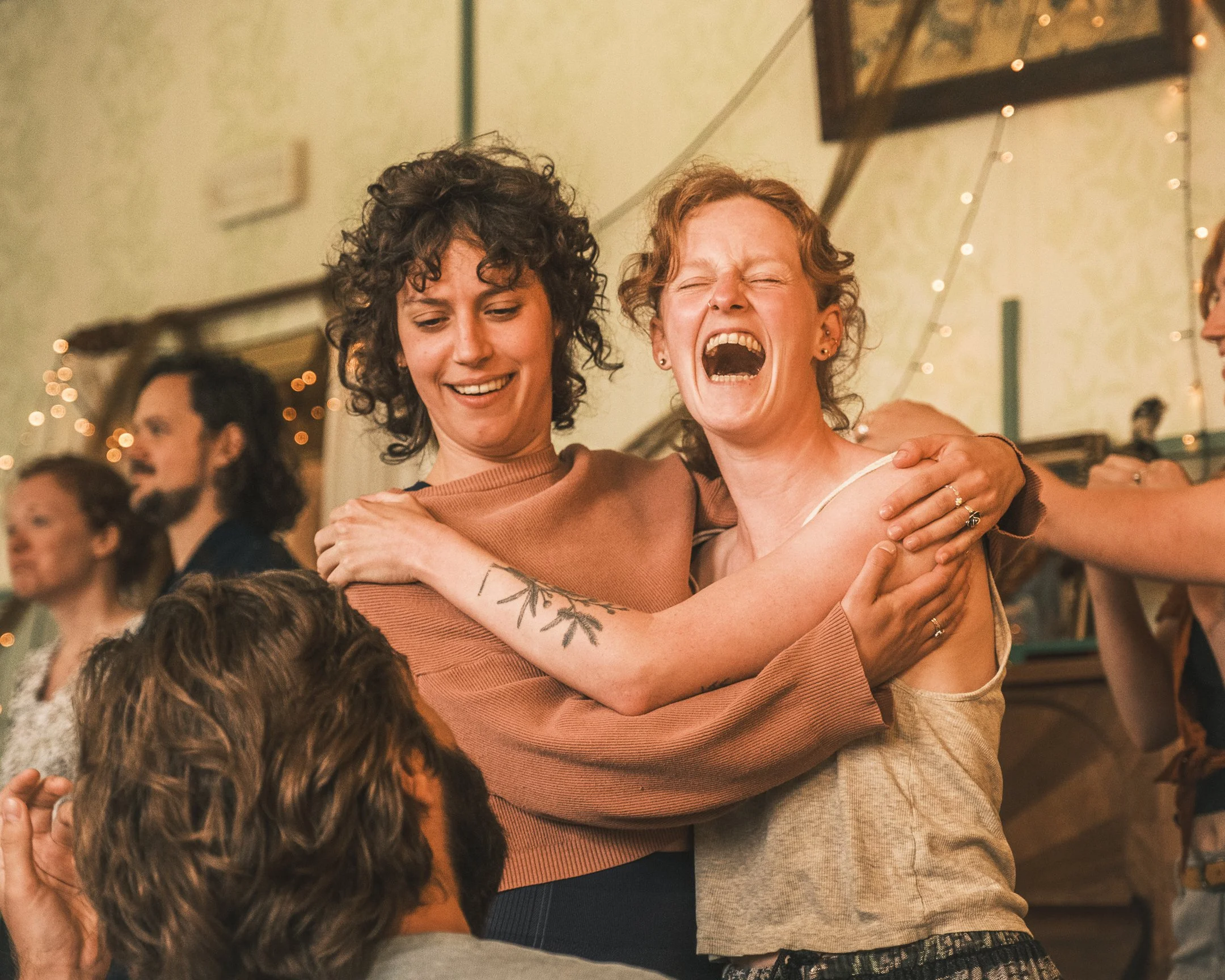 Twee vrouwen omhelzen en lachen op een feest, met andere gasten op de achtergrond.