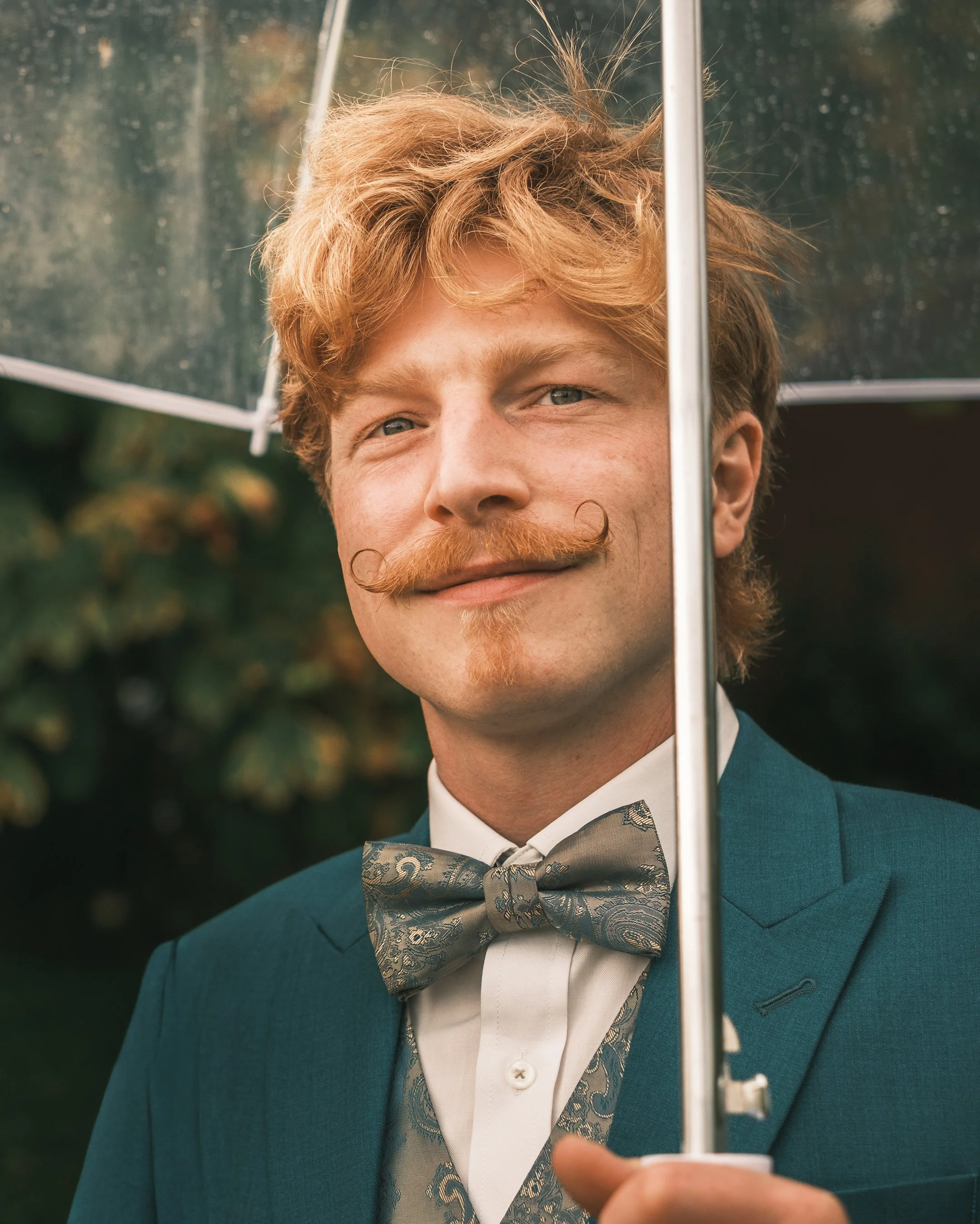 A man with red hair, a handlebar mustache, and a goatee smiles while holding a transparent umbrella in front of him. He is wearing a teal suit and a patterned bow tie. The background is leafy and green.