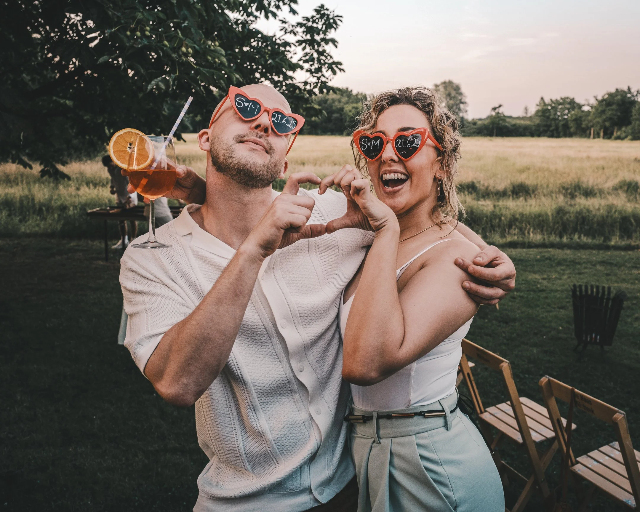 Twee mensen maken een hartvorm met hun handen, dragen hartvormige zonnebrillen met datums en schrijven 'SM' op de brillen, terwijl ze samen lachen in een buitenomgeving.