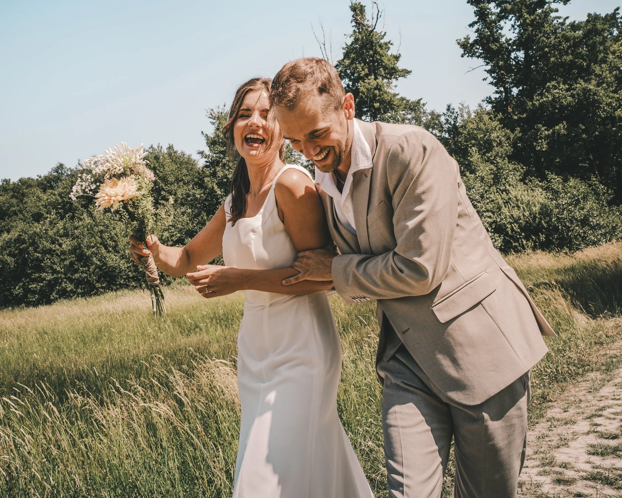 Trouwende paar in een veld, de bruid houdt een boeket, de bruidegom glimlacht terwijl hij haar vasthoudt, beide lachen.