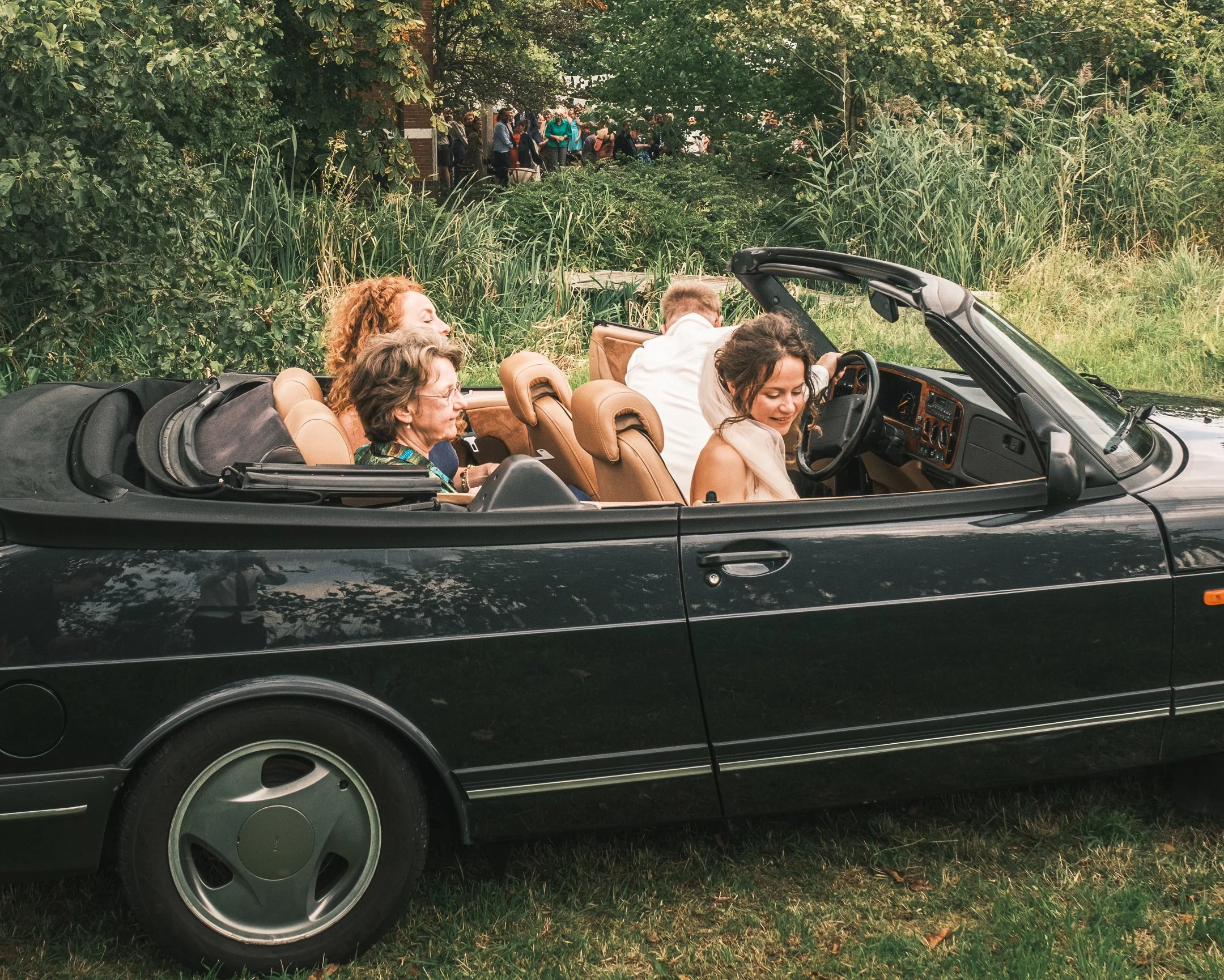 Een groep mensen in een zwarte cabriolet met open dak, rijdend langs een groene natuuromgeving.