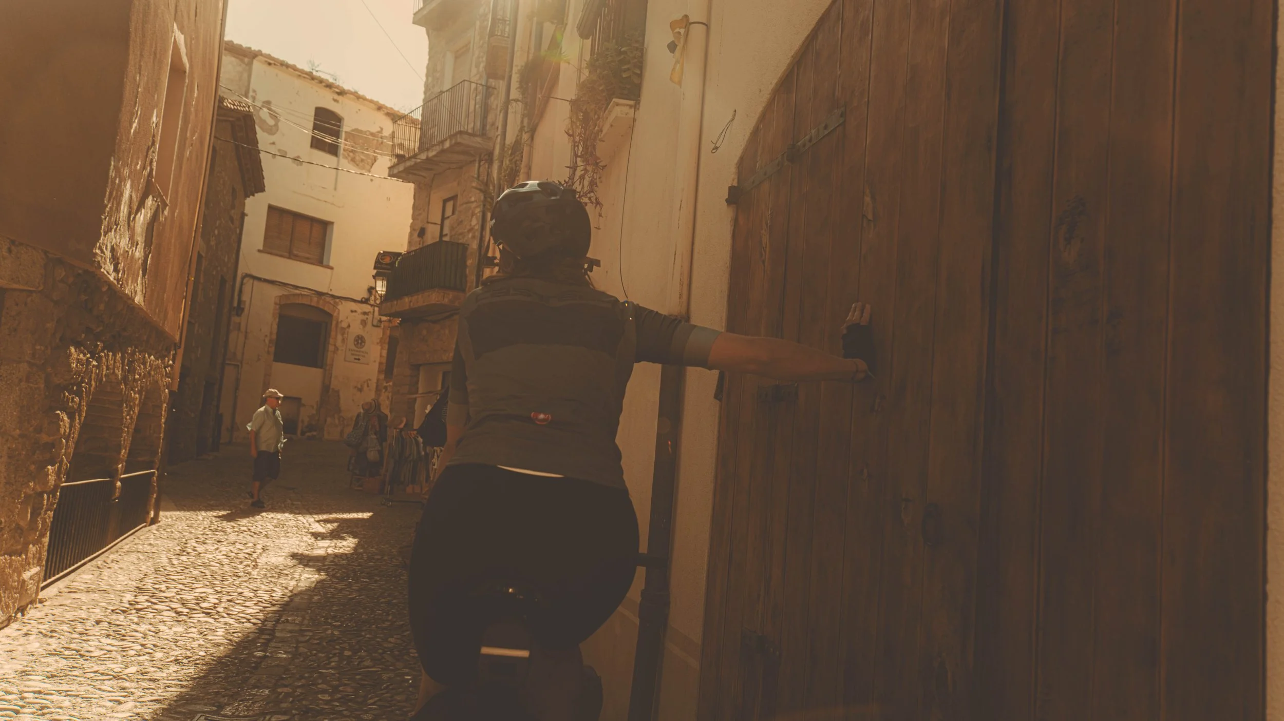 Persoon met helm en rugzak die een grote houten deur op een smal geplaveid straatje duwt, met enkele mensen op de achtergrond.