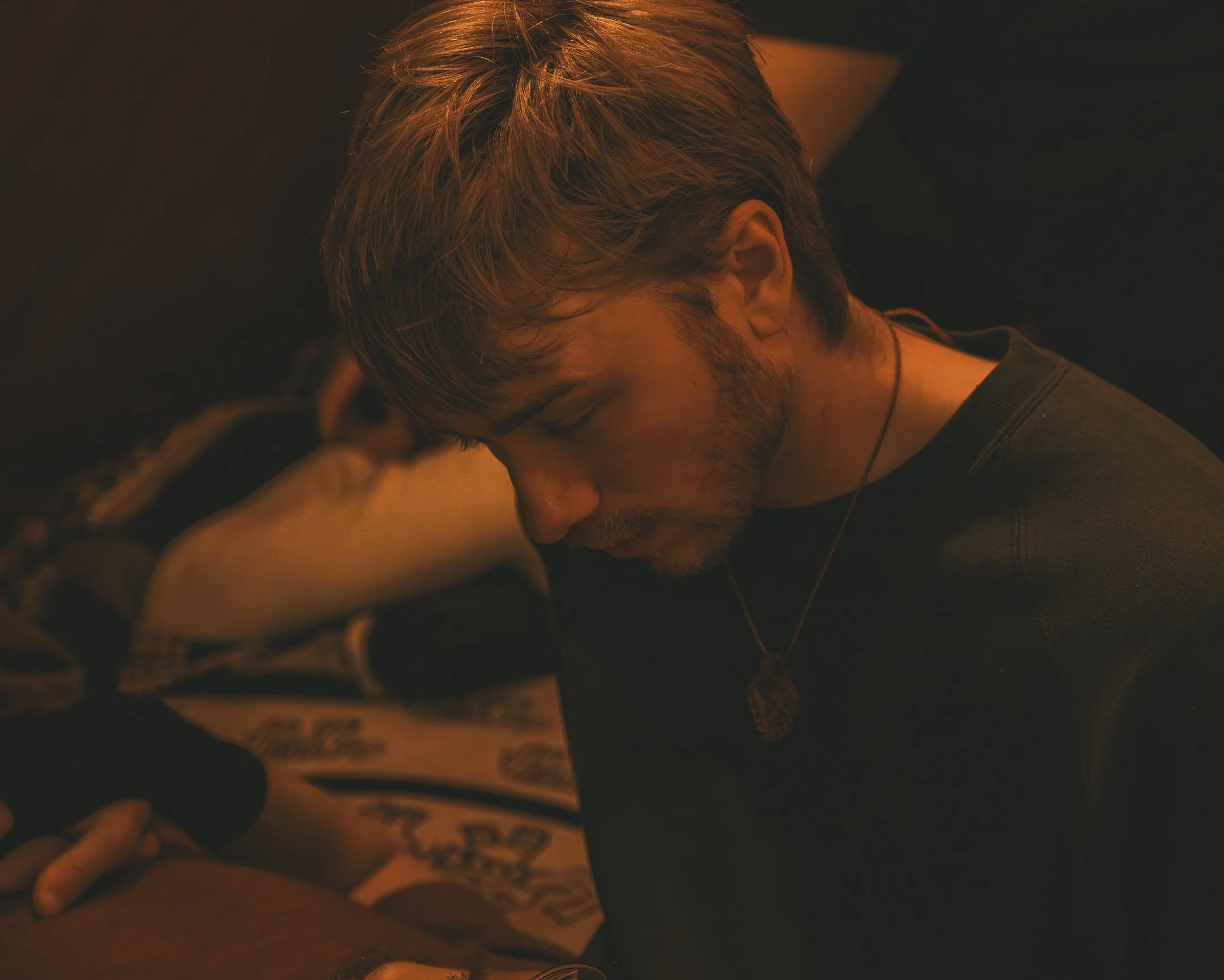 Een jonge man met bruin haar en een baard, met ogen dicht, leunend op een tafel of oppervlak, onder een warme oranje gloed, met een medailles aan een ketting om de hals.