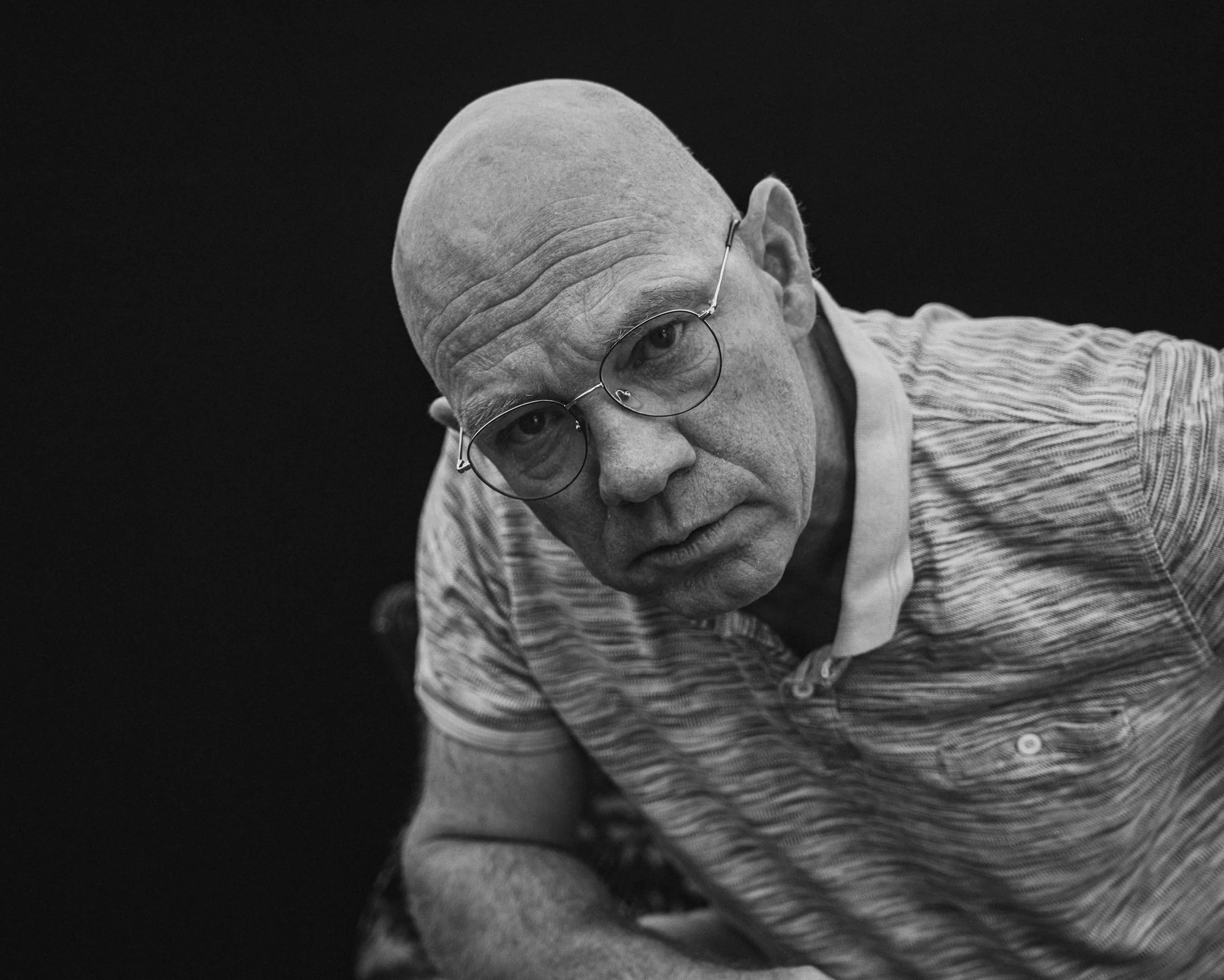 Zwart-wit close-up foto van een volwassen man met een kale hoofd, bril, en een gestreept polo shirt, met een neutrale uitdrukking, tegen een donkere achtergrond.