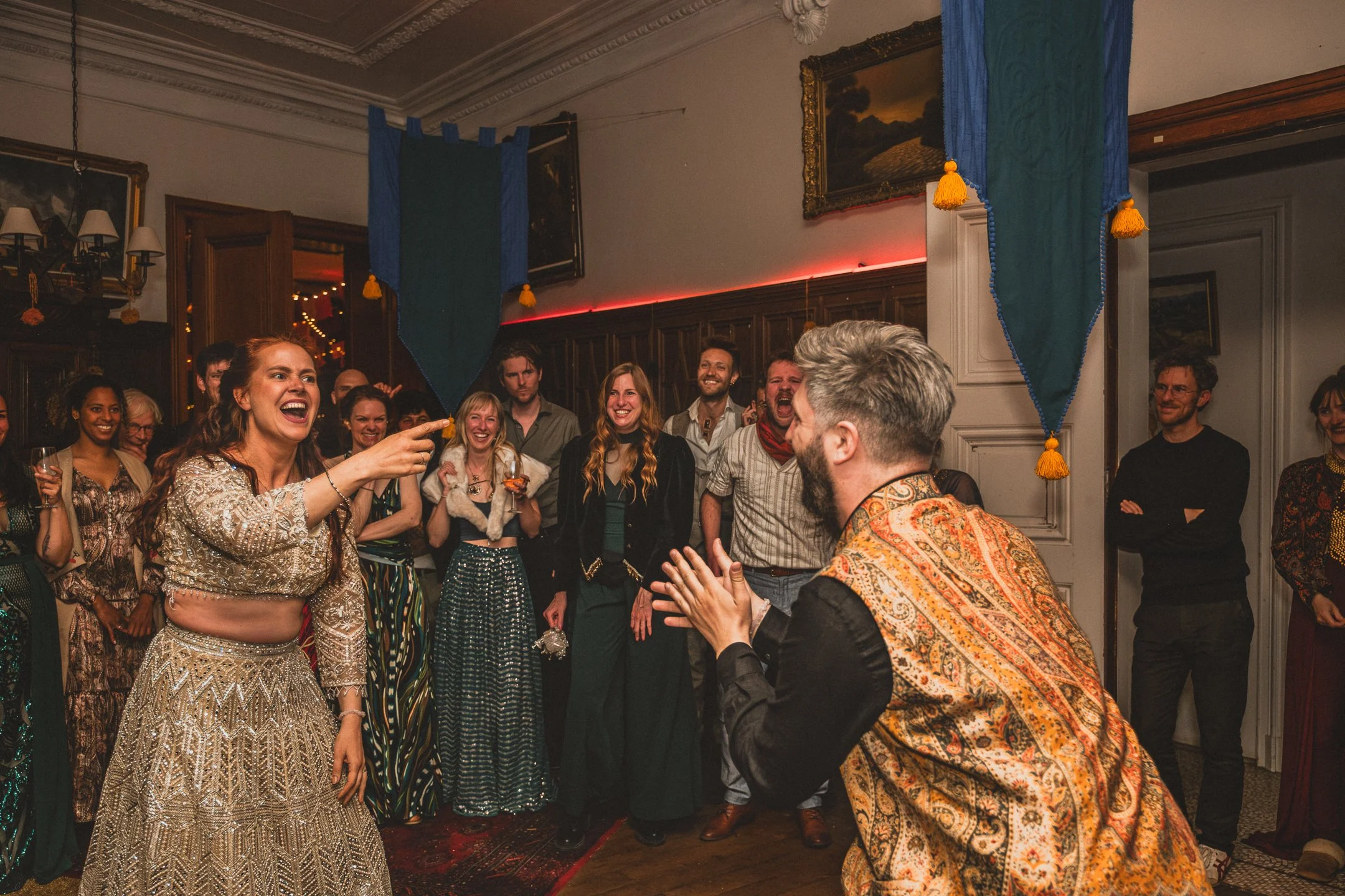 Mensen die lachen en plezier maken tijdens een feest in een stijlvolle, klassieke kamer met donkerhouten panelen, schilderijen en blauwe vlaggen met gele franjes.