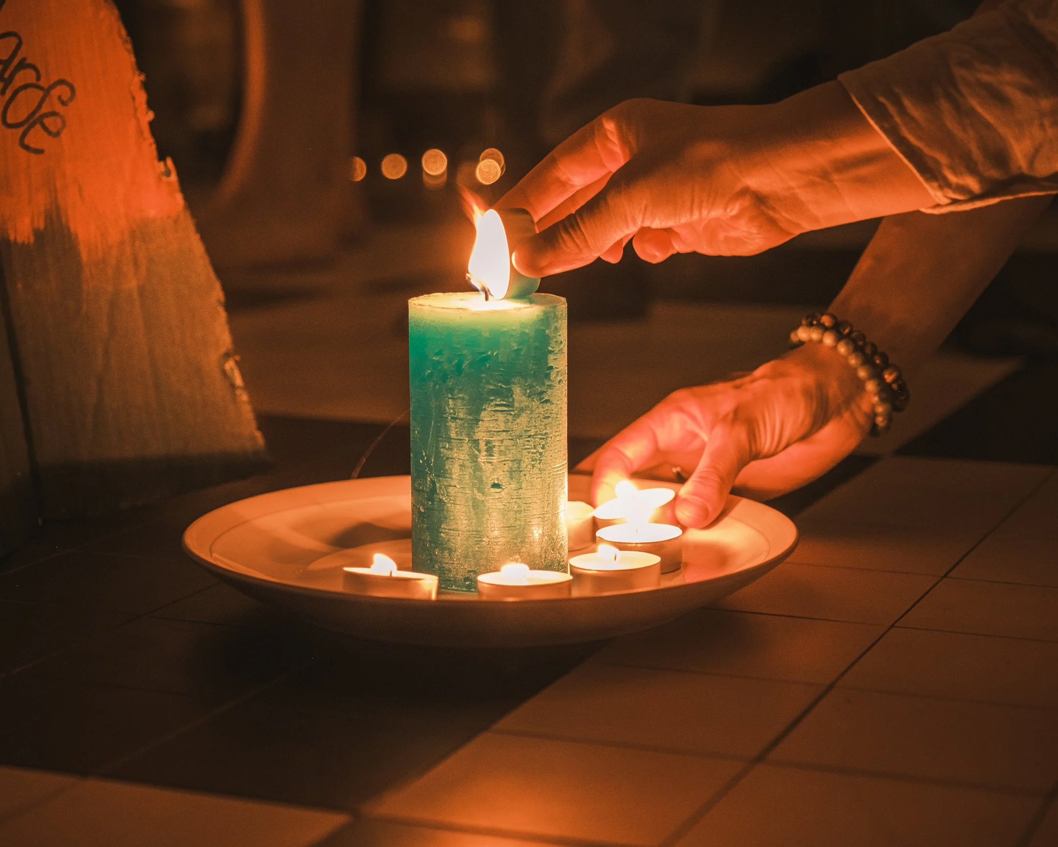 Person die een grote groene kaars aansteekt die op een dienblad ligt met meerdere kleine waxinelichtjes eromheen, in een dimverlichte omgeving.