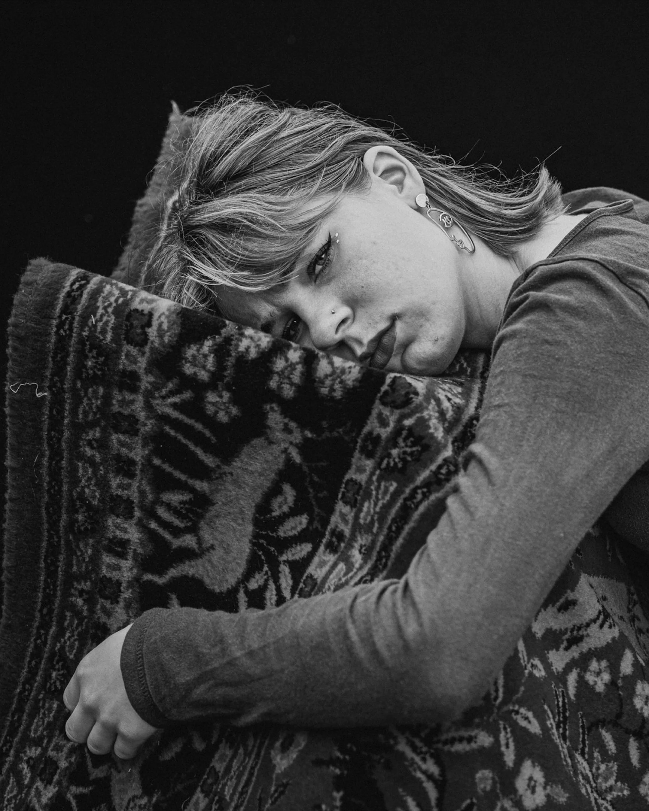 Zwarte en witte foto van een jonge vrouw die op een gestreepte deken ligt, haar hoofd rustend op kussen, met blonde haar, sieraden, en een serieuze uitdrukking.