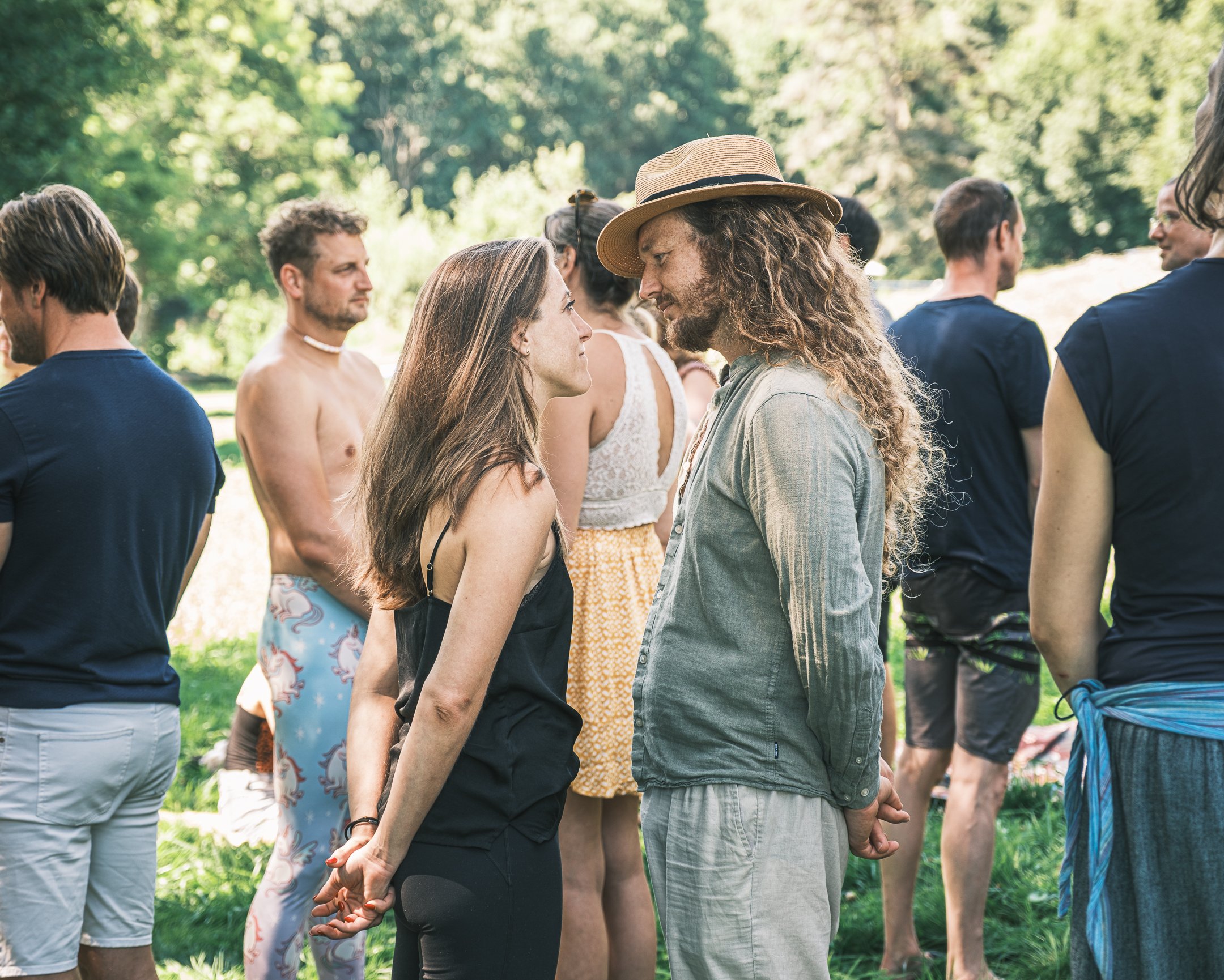A group of people outdoors in a park, with two individuals in the center facing each other, holding hands behind their backs, about to kiss. Others are engaged in conversations or standing nearby.