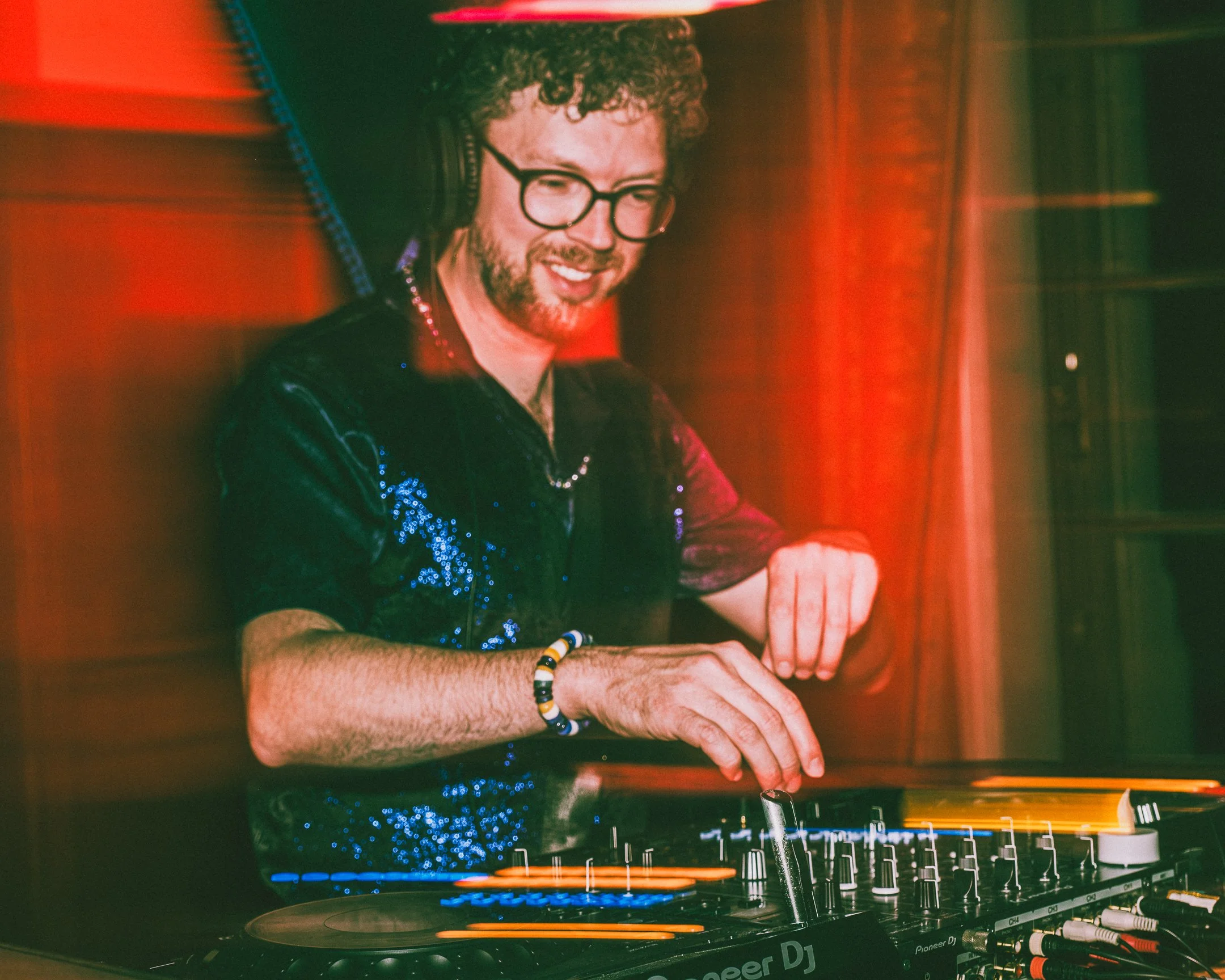 Een man met bril en een bearmuts draait muziek op een DJ-overslag, omgeven door een kleurrijke, lichte achtergrond.