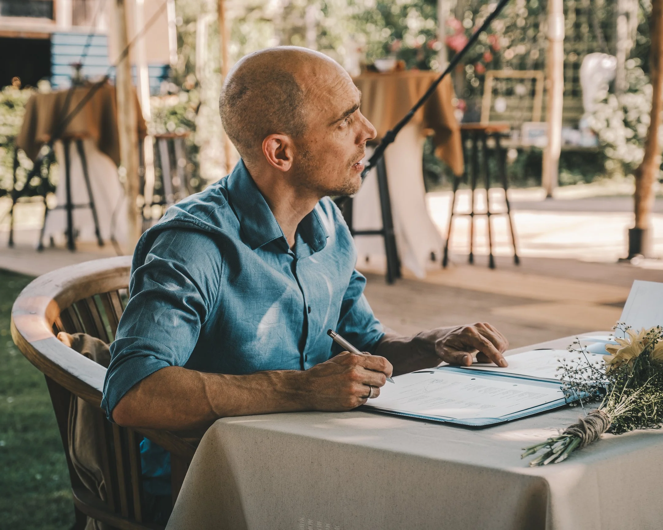 Man zit aan een tafel met een boek en een pen, op een houten stoel, mogelijk in een buitenruimte als een tuin met natuurlijke lichteffecten.