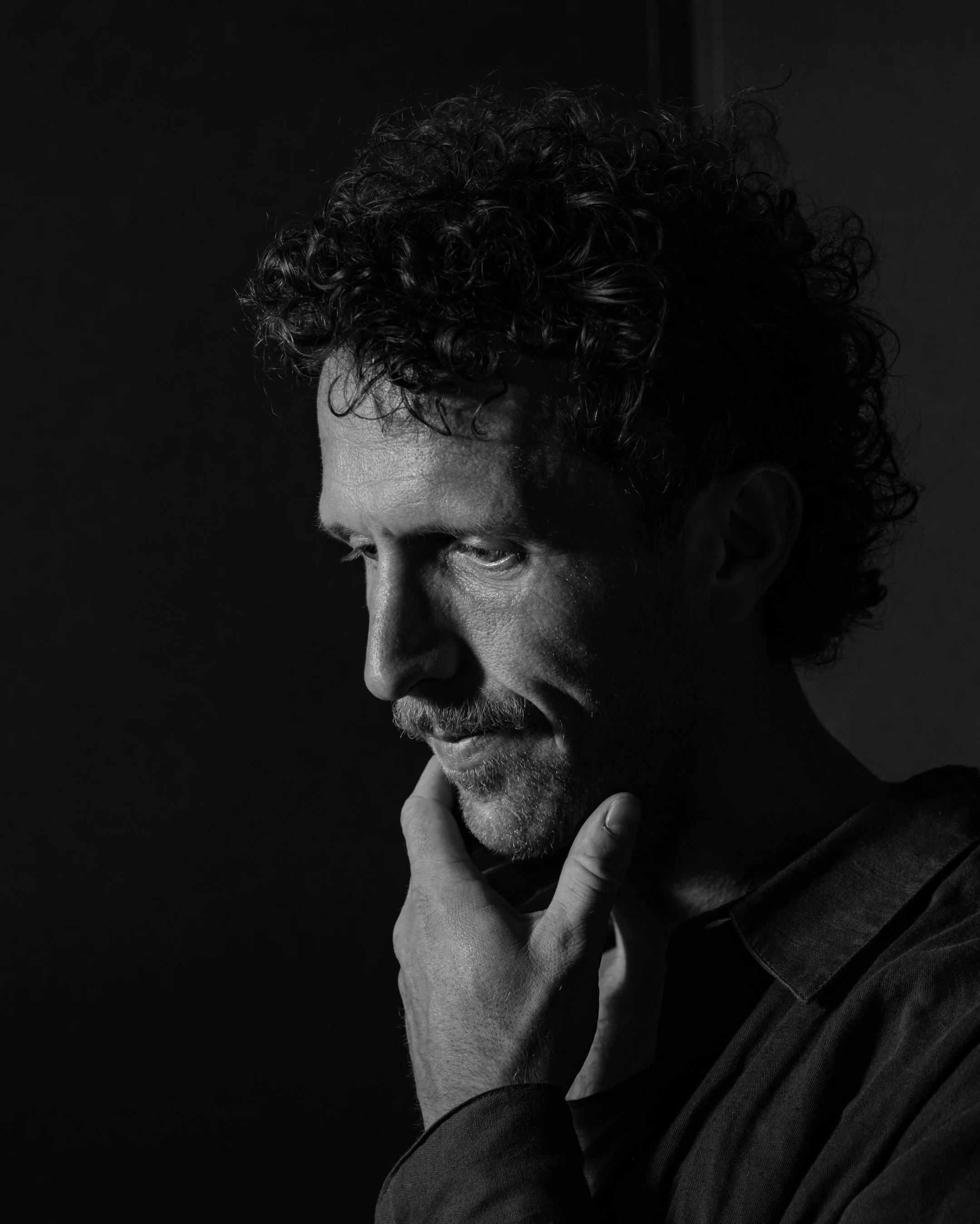 Zwarte en witte foto van een man met halflang, gekruld haar en een baard, die nadenkt met zijn hand op zijn kin tegen een donkere achtergrond.