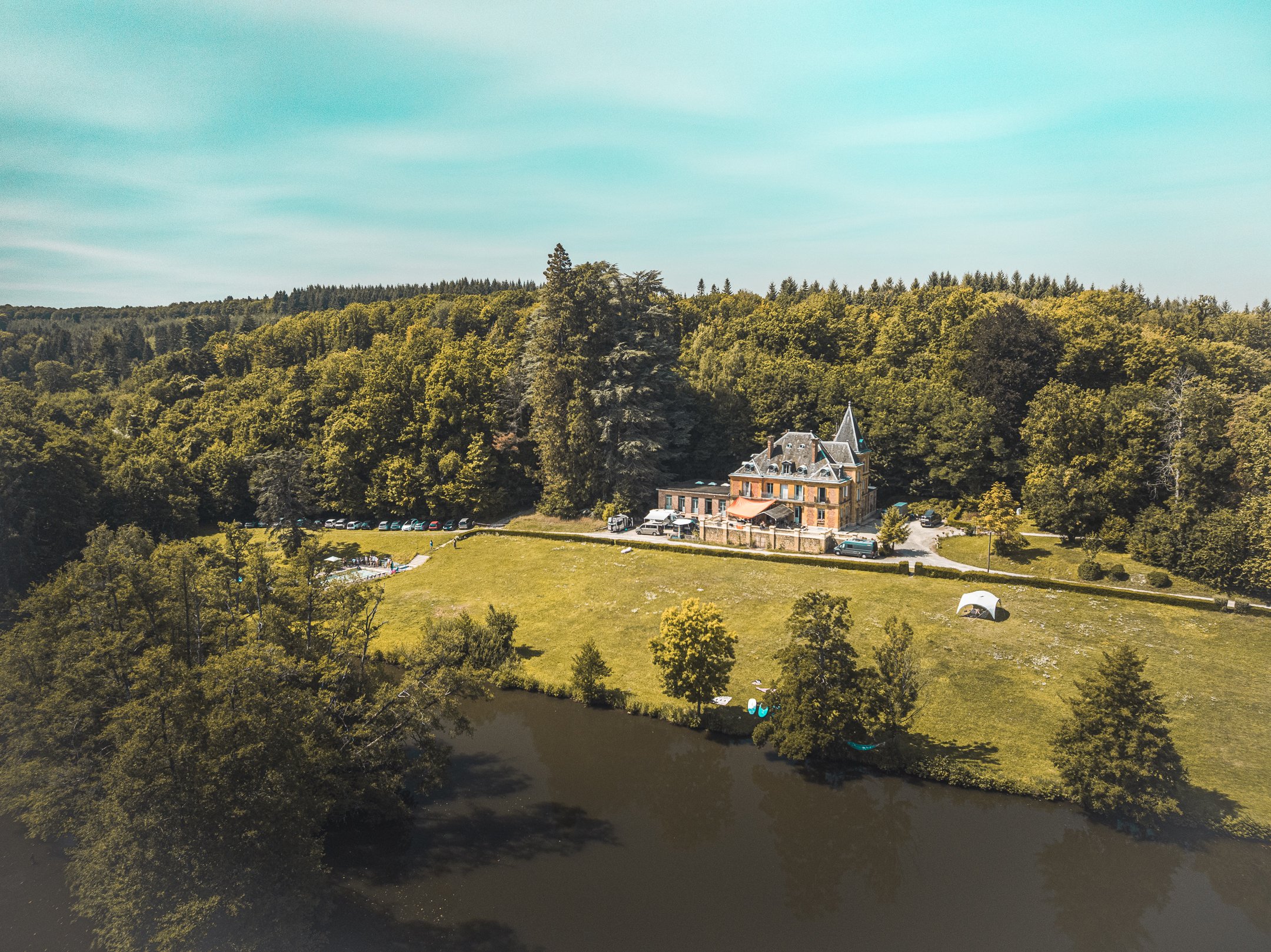 Een groot, kasteelachtig huis met een toren, omgeven door een bos en een klein meer op de voorgrond, met een grasveld en enkele bomen.