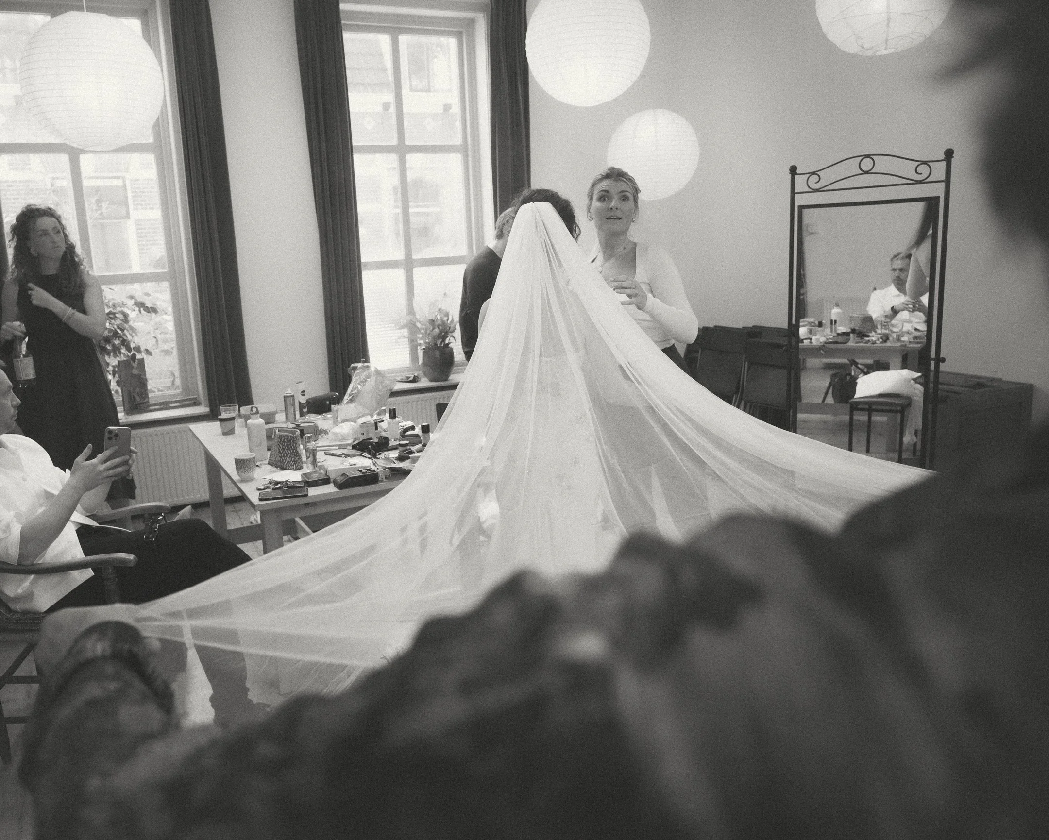 Vrouw in witte jurk met lange sluier wordt geholpen door vriendinnen en stylisten in een verlichte kamer met grote ramen en ronde lampen