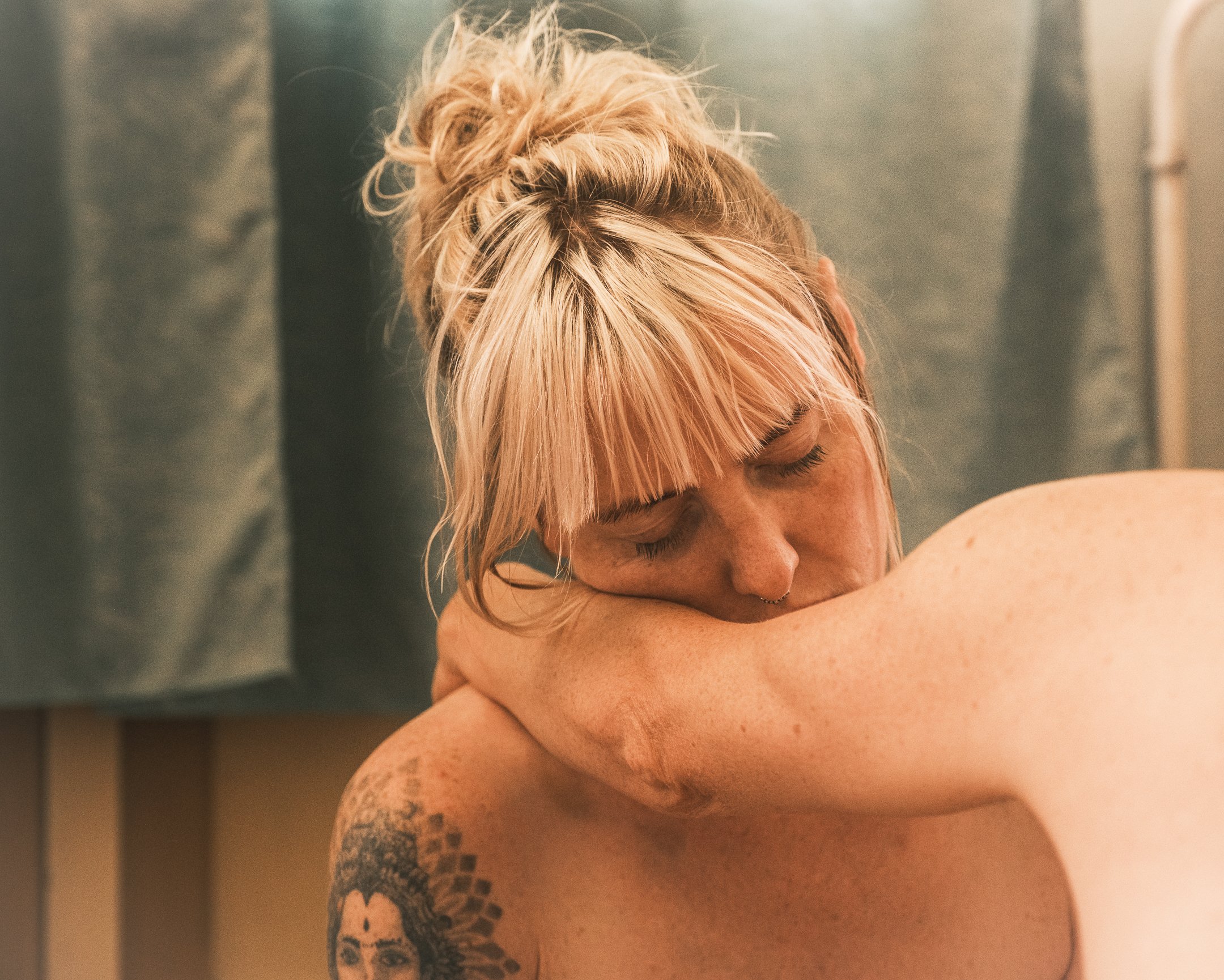 A woman with blonde hair in a messy bun, with bangs covering her forehead, resting her forehead on her arm, eyes closed, showing a tattoo of a woman's face with a floral design on her shoulder.