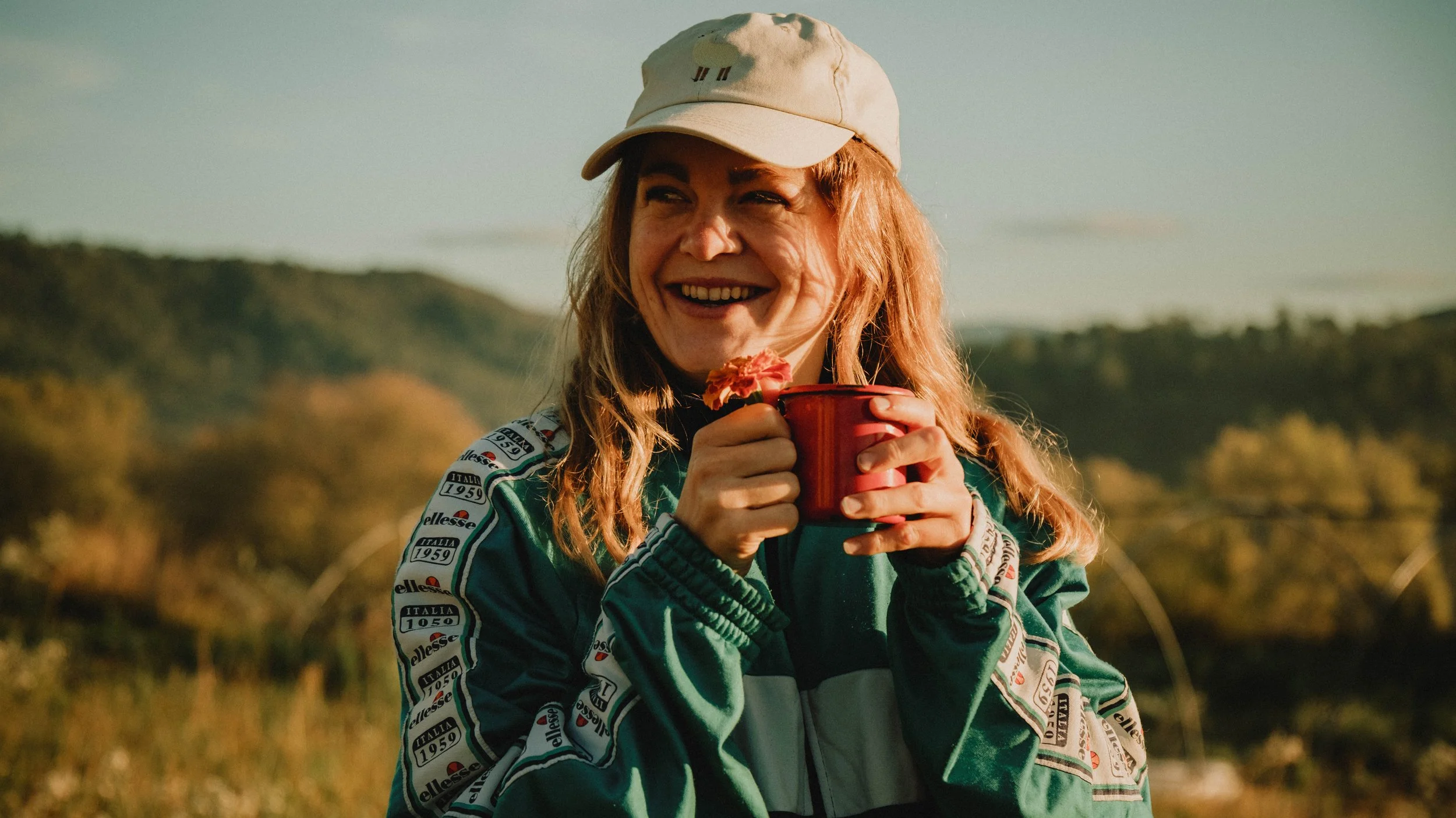 Vrouw met rood haar, lachend, draagt een witte pet en groene jas, houdt een rode mok met bloemen tegen haar gezicht, buiten met heuvels op de achtergrond.