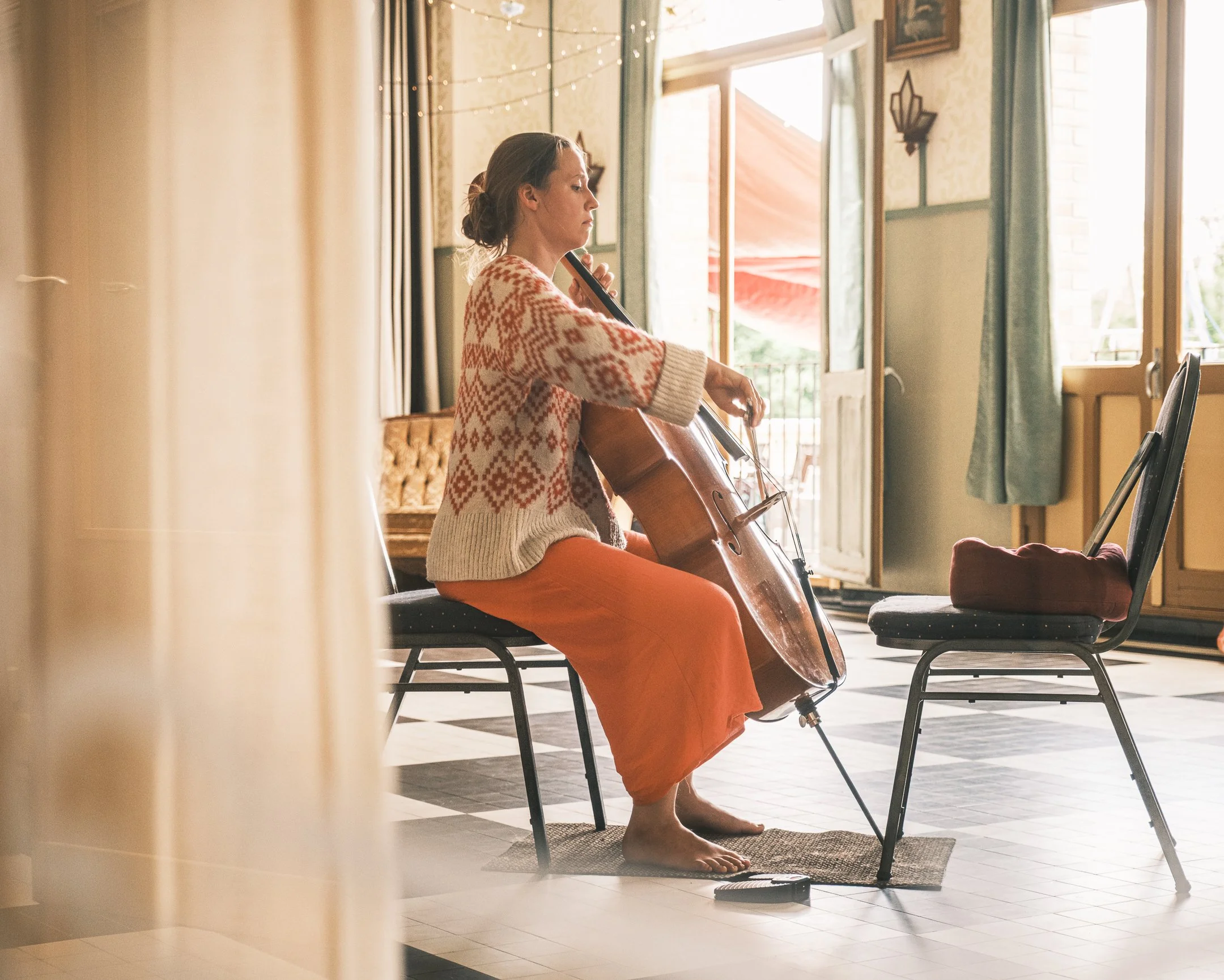 Woman sitting barefoot on a chair, playing a cello in a sunlit room with open doors, curtains, and wall decorations.