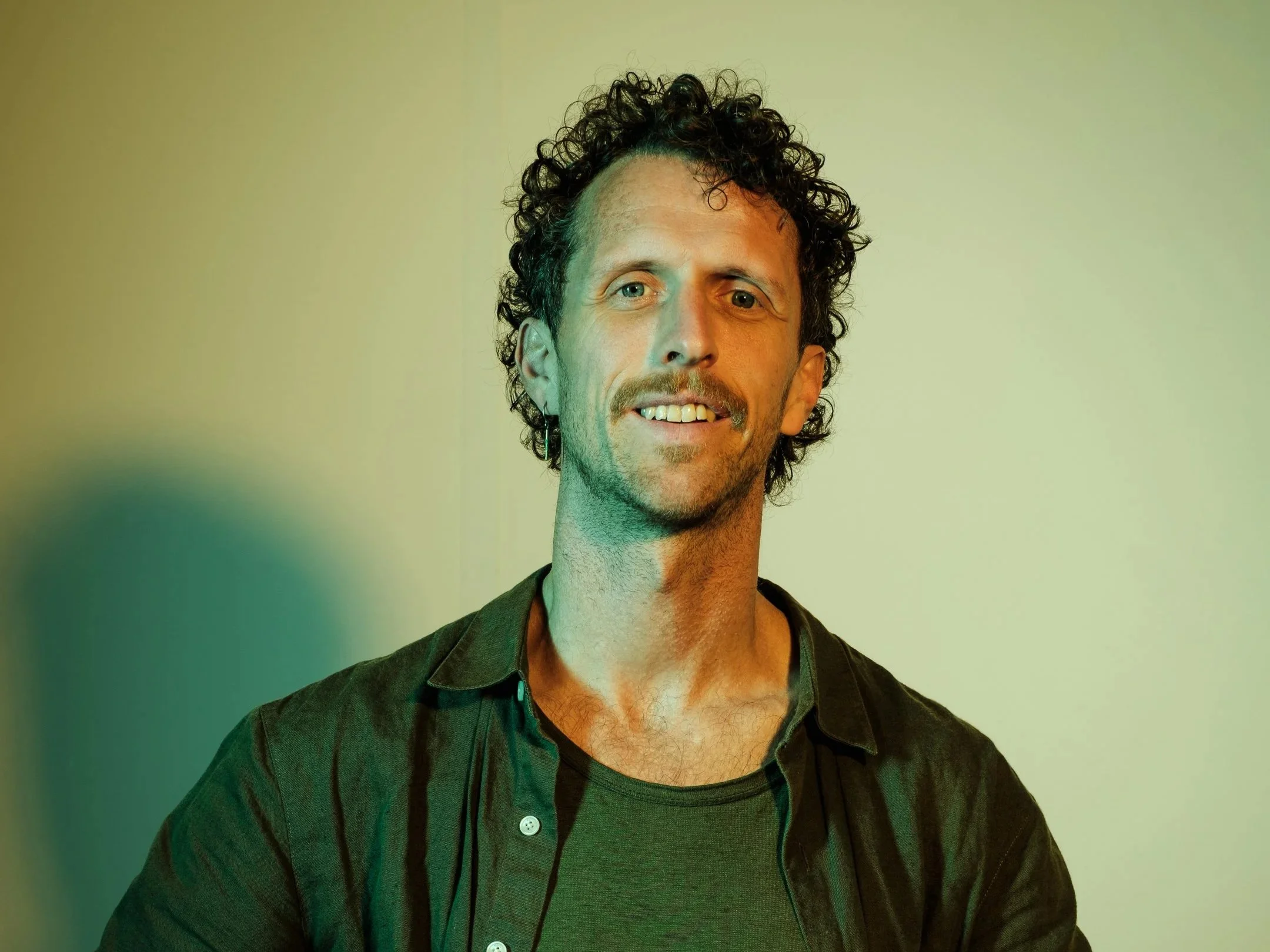 Portret van een man met curly haar die lacht, tegen een lichte achtergrond.