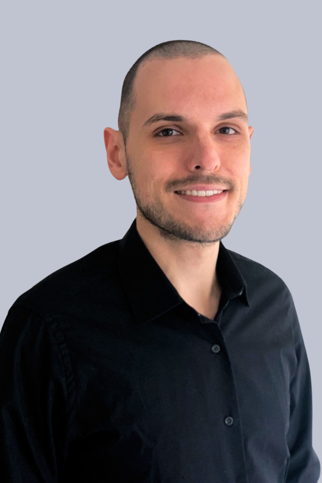 Retrato de un hombre joven con cabello rapado, barba corta, sonriendo, usando una camisa negra, fondo de color gris claro.