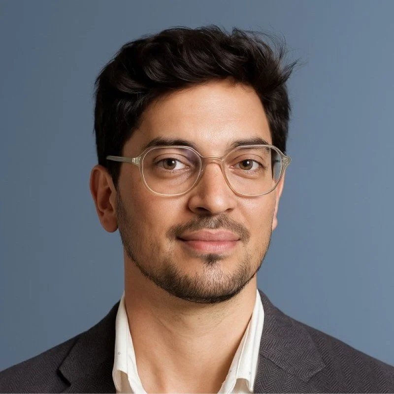 Retrato de un hombre con gafas transparentes, cabello oscuro, barba y bigote cortos, vestido con saco gris y camisa blanca, sonriendo ligeramente con fondo azul.