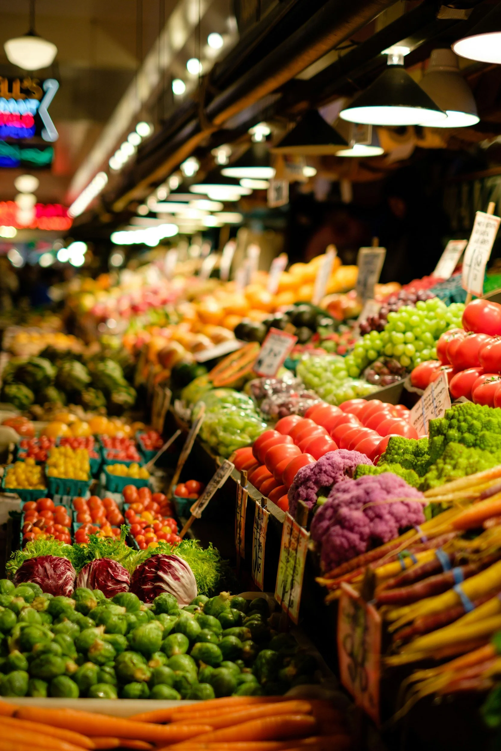Colorful display of fresh vegetables and fruits at an outdoor market, including tomatoes, cauliflower, Brussels sprouts, and carrots.