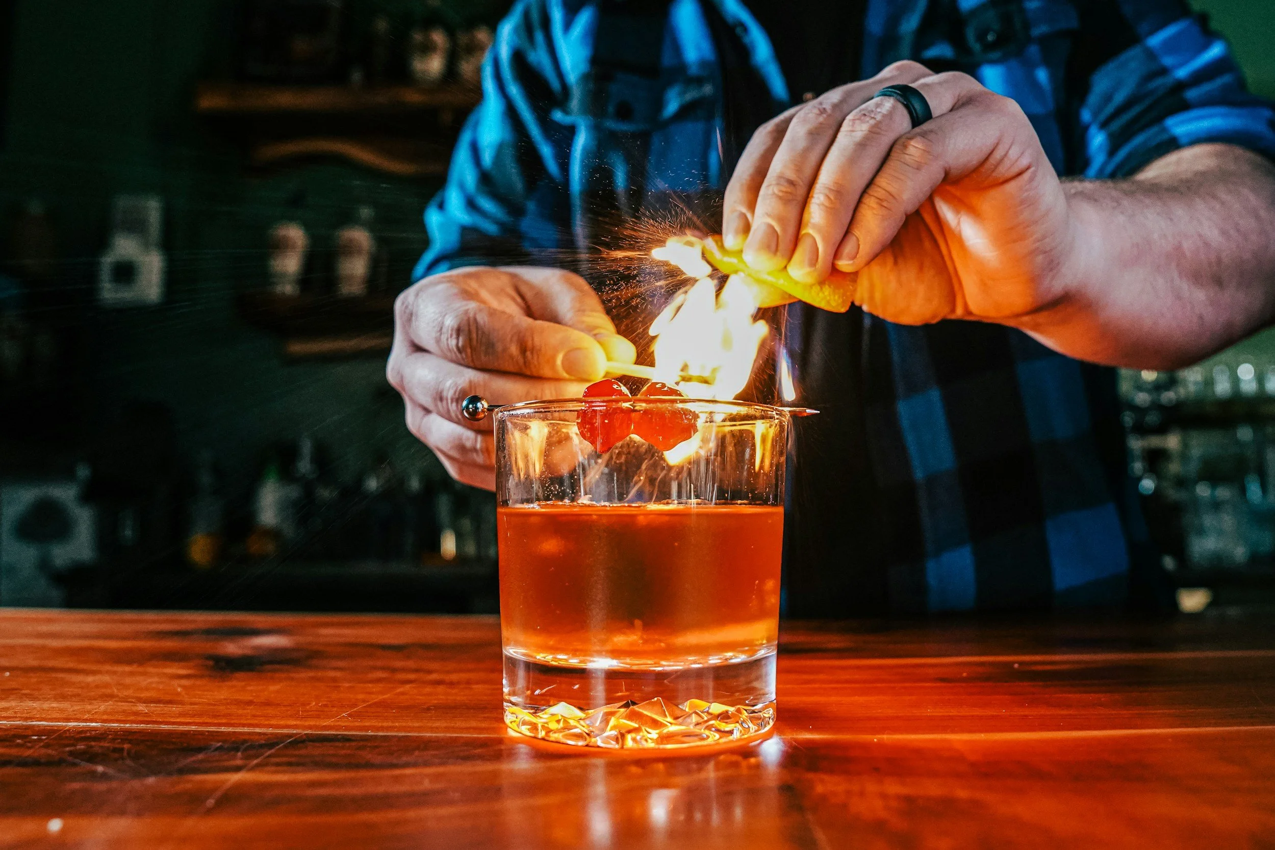A person in a blue plaid shirt is lighting a cherry garnish with a flame on a cocktail in a glass on a wooden bar counter.