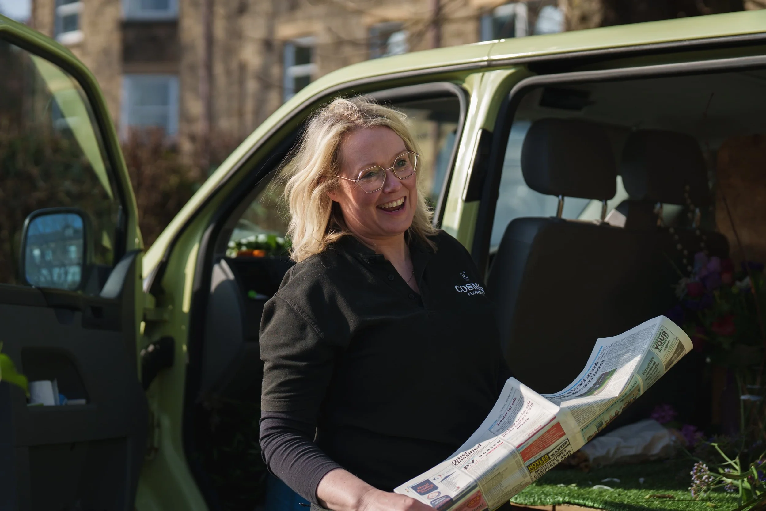 Woman with blonde hair and glasses smiling, standing next to an open green vehicle, holding a large newspaper or flyer.