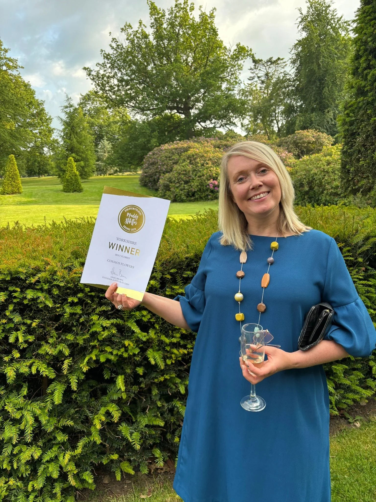 Woman in a blue dress holding a certificate and a glass of champagne in a garden.
