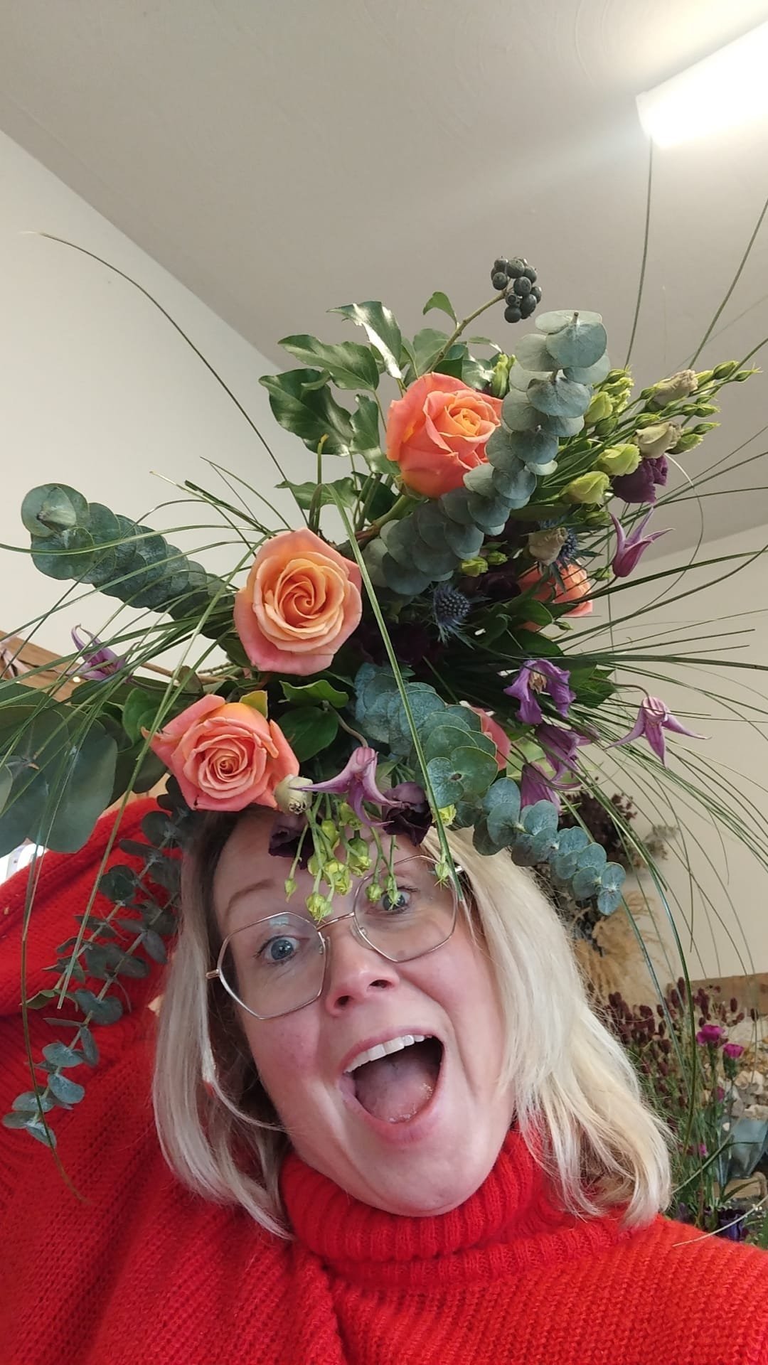 Woman with glasses wearing a red sweater, holding a large flower bouquet above her head, with an excited expression.