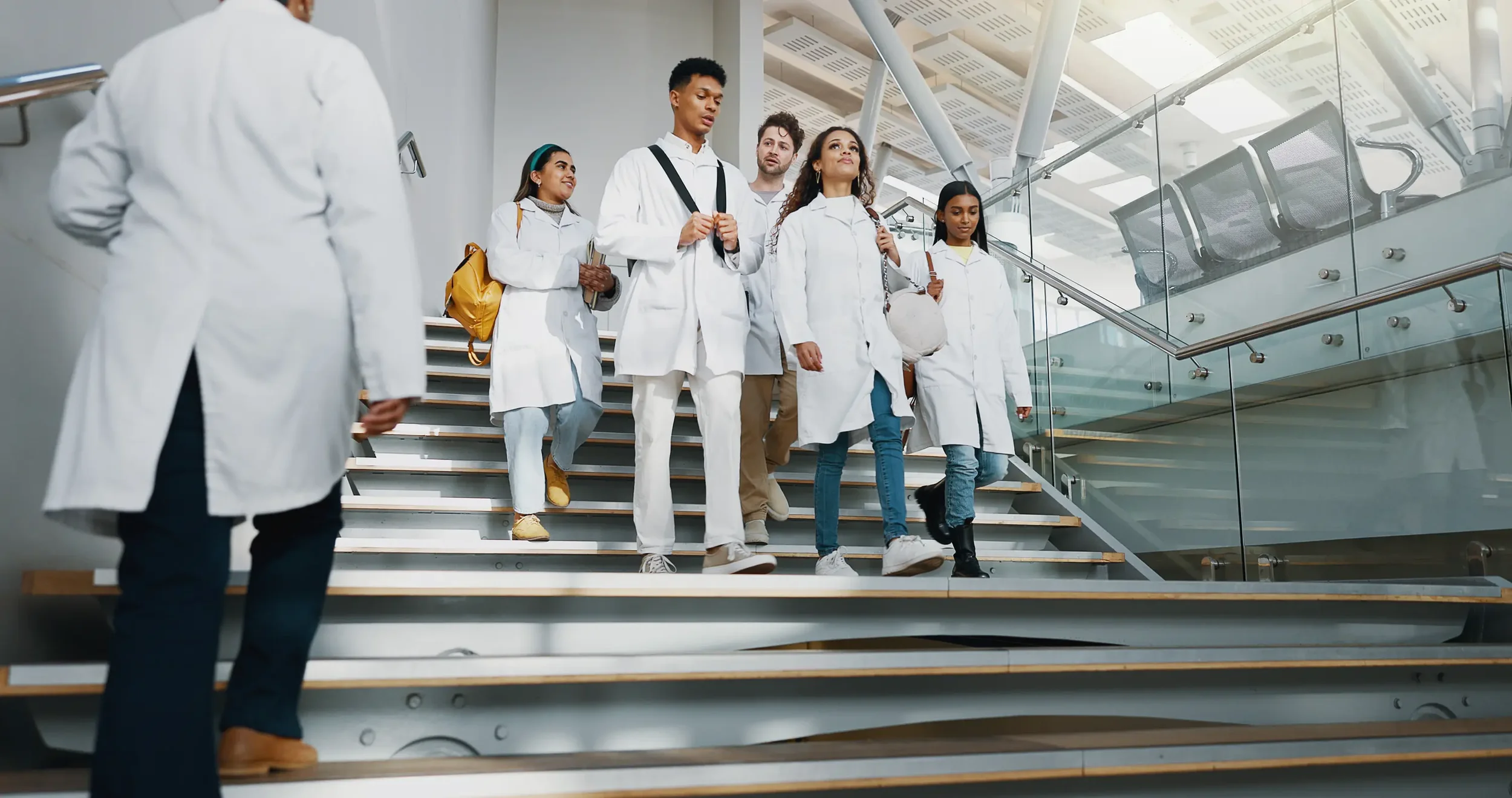 Group of diverse medical students in white lab coats walking down stairs in a modern hospital or medical center.