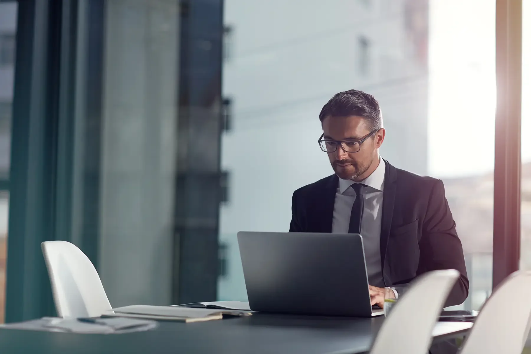 Businessman in a formal suit working on a laptop in a modern office with large windows.