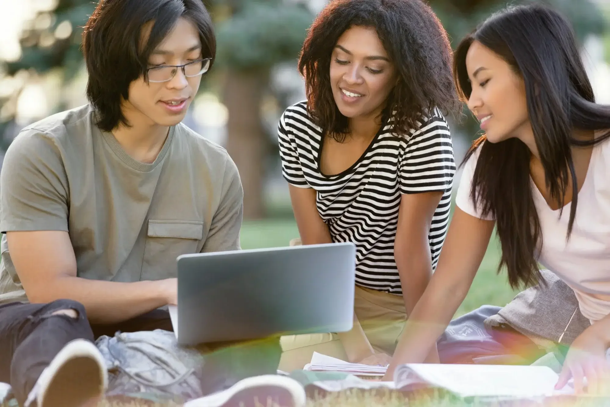 Three young people sitting outdoors on grass, looking at a laptop together and smiling.
