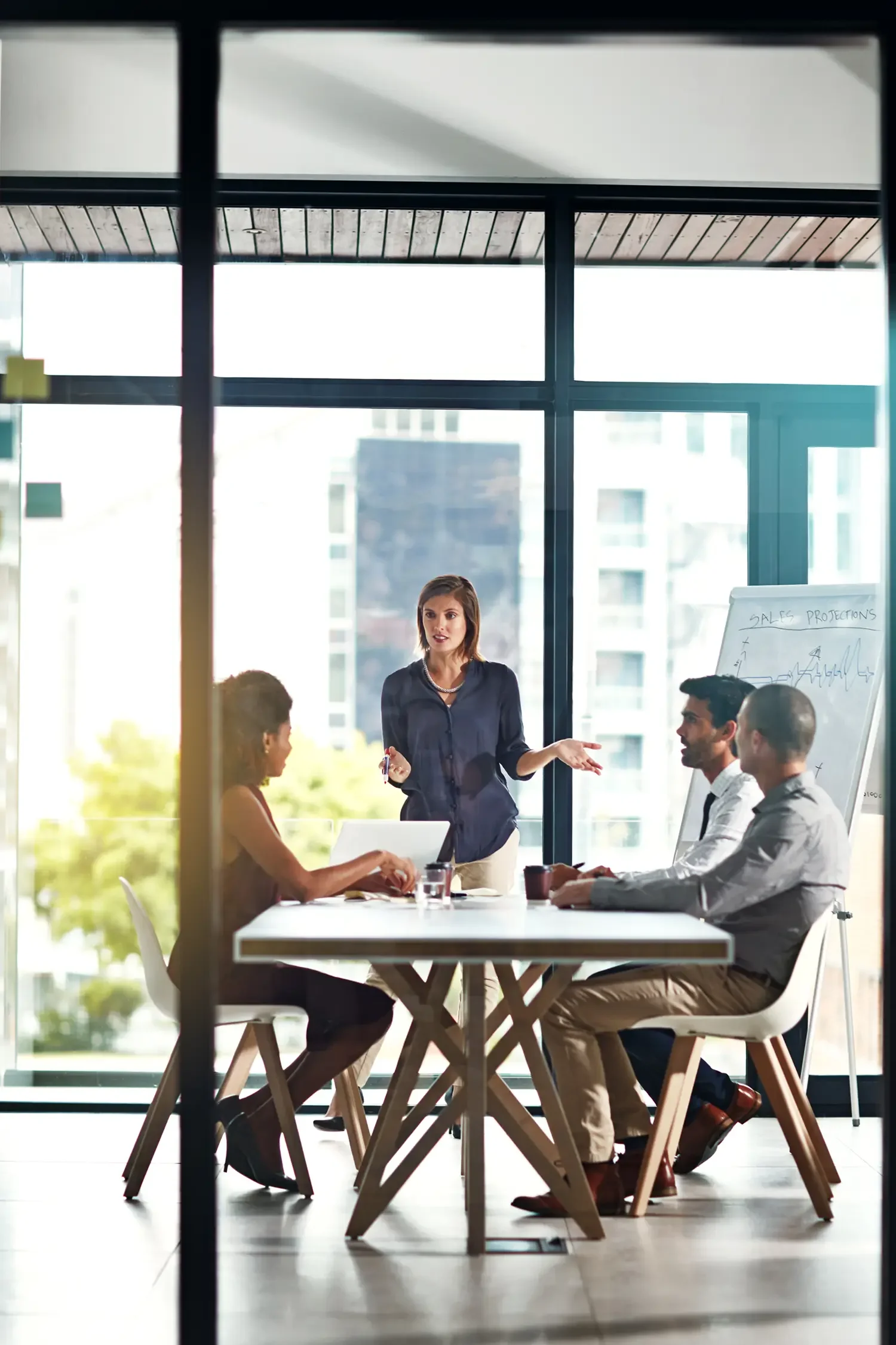Businesswoman giving a presentation in a modern office, with three colleagues seated at a table, a large window with a city view in the background.