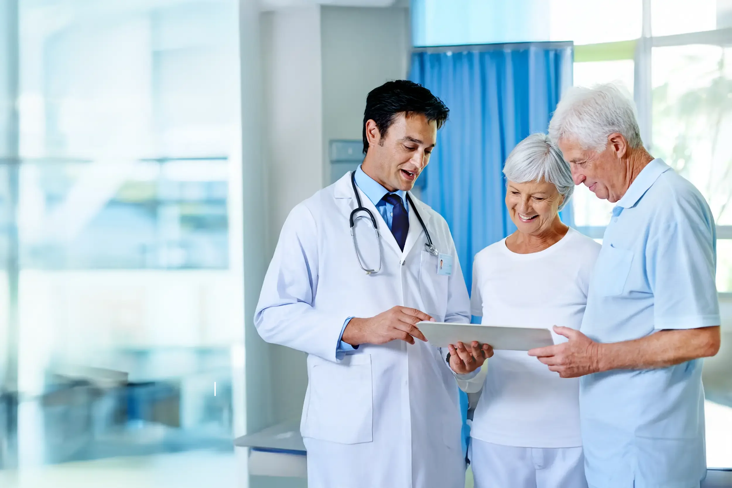 Two elderly patients and one doctor, looking at a tablet together in a hospital room with large windows and blue curtains.