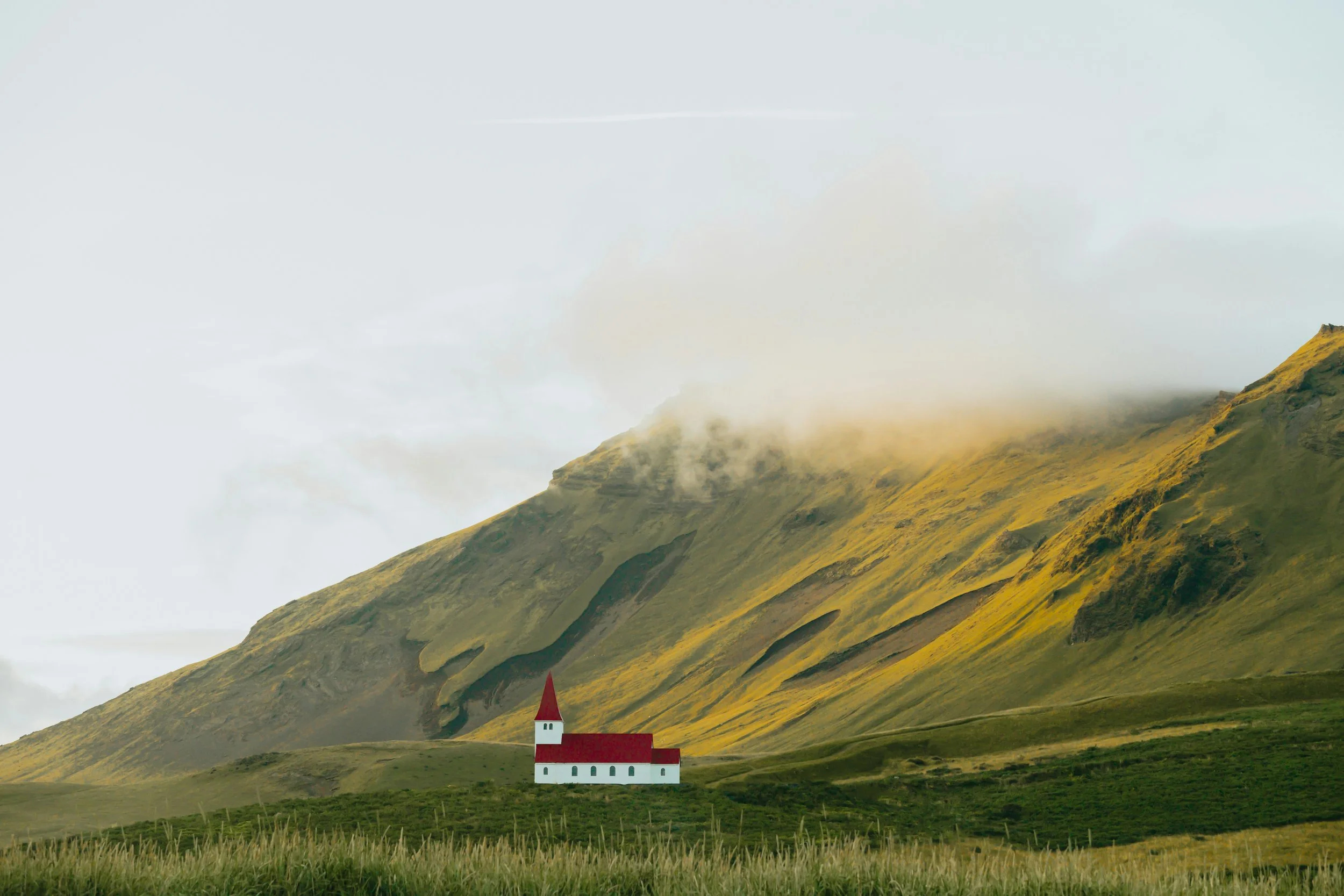 A small white church with a red roof and steeple situated on green grassy land with a large mountain in the background, partly covered in clouds and fog.
