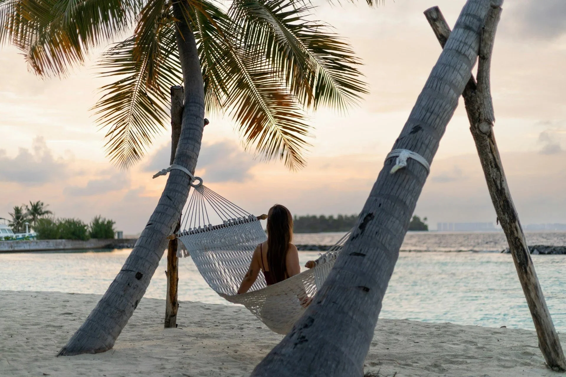 A woman sitting on a hammock tied between two palm trees on a sandy beach, watching the sunset over the water.