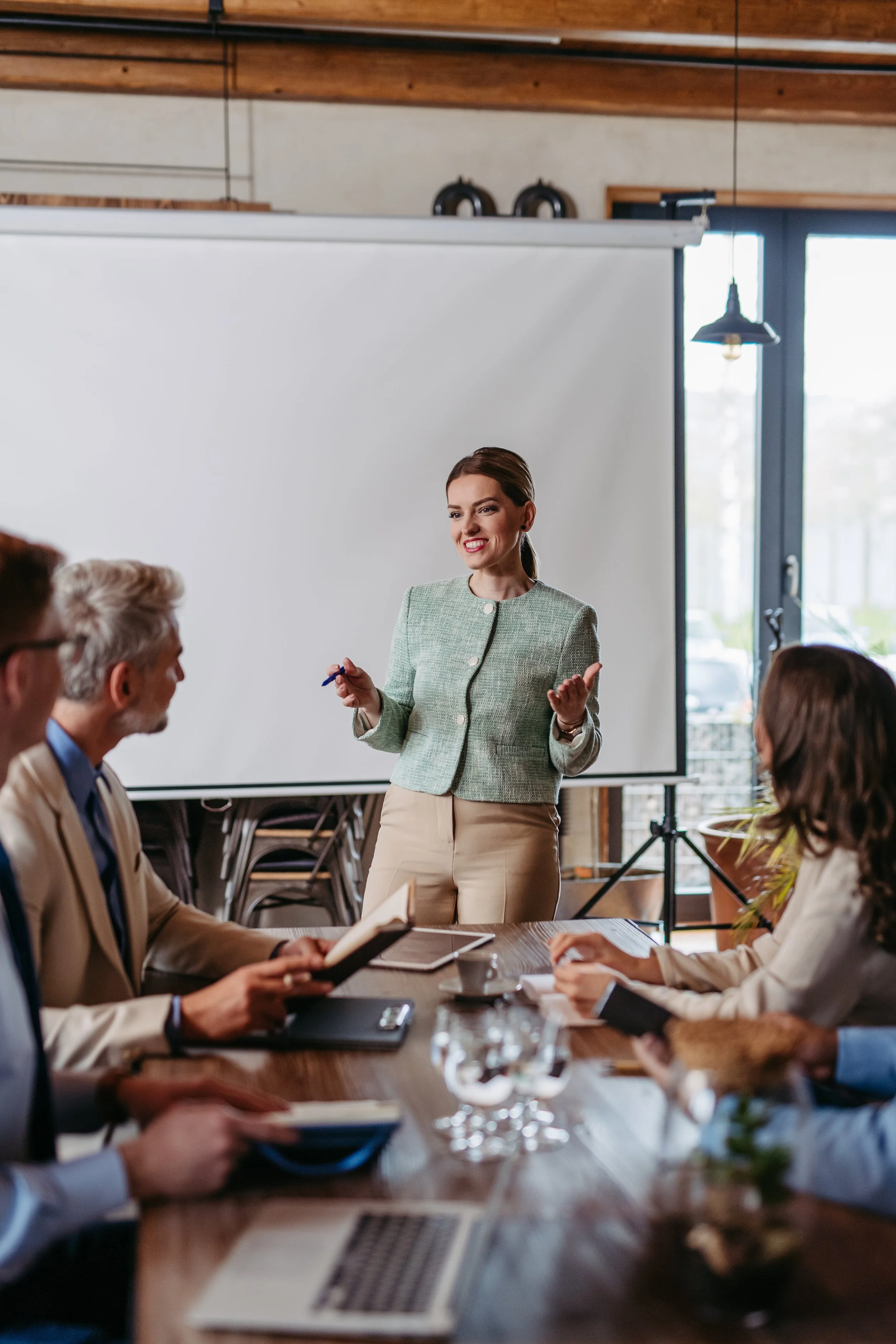 A woman in a green blazer and beige pants giving a presentation to a group of professionals in a conference room using the book rollout as helpful too for implementing eos to the organization