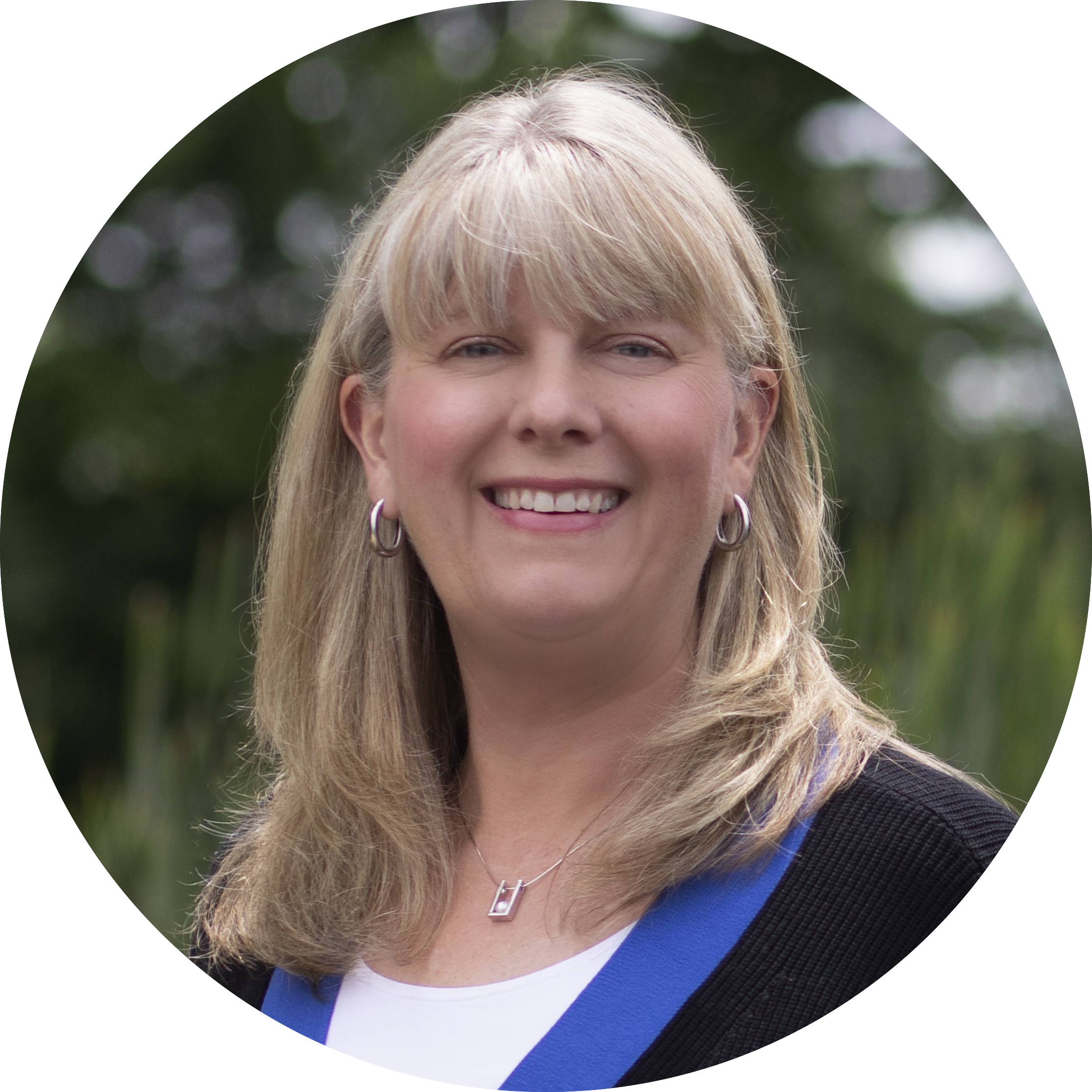 Expert EOS implementer Marisa Smith with shoulder-length blonde hair wearing silver hoop earrings, a silver necklace, a white top, and a dark jacket, smiling outdoors with green foliage in the background.