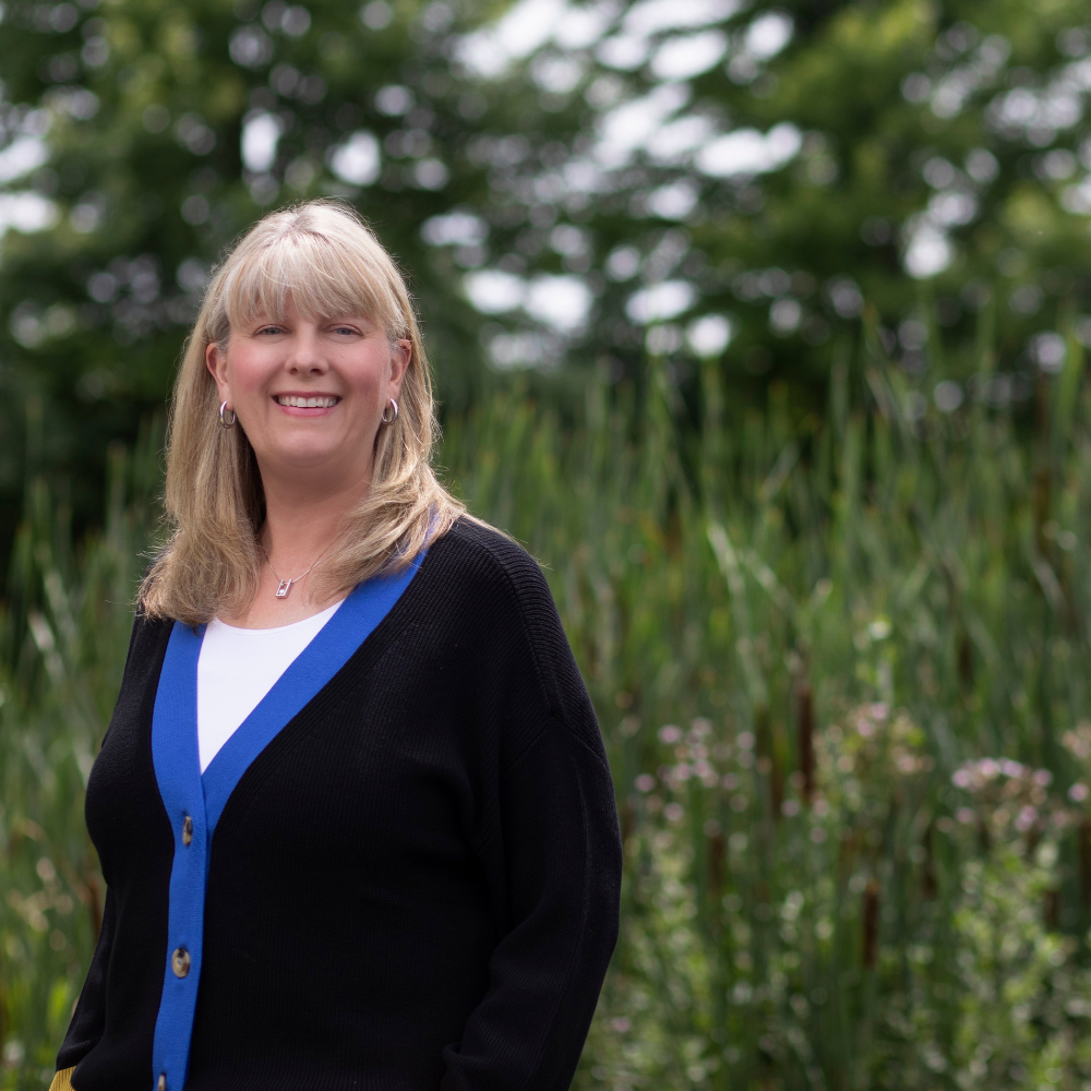 expert eos implementer marisa smith with blonde hair wearing a black cardigan with blue trim, standing outdoors with green trees and foliage in the background.