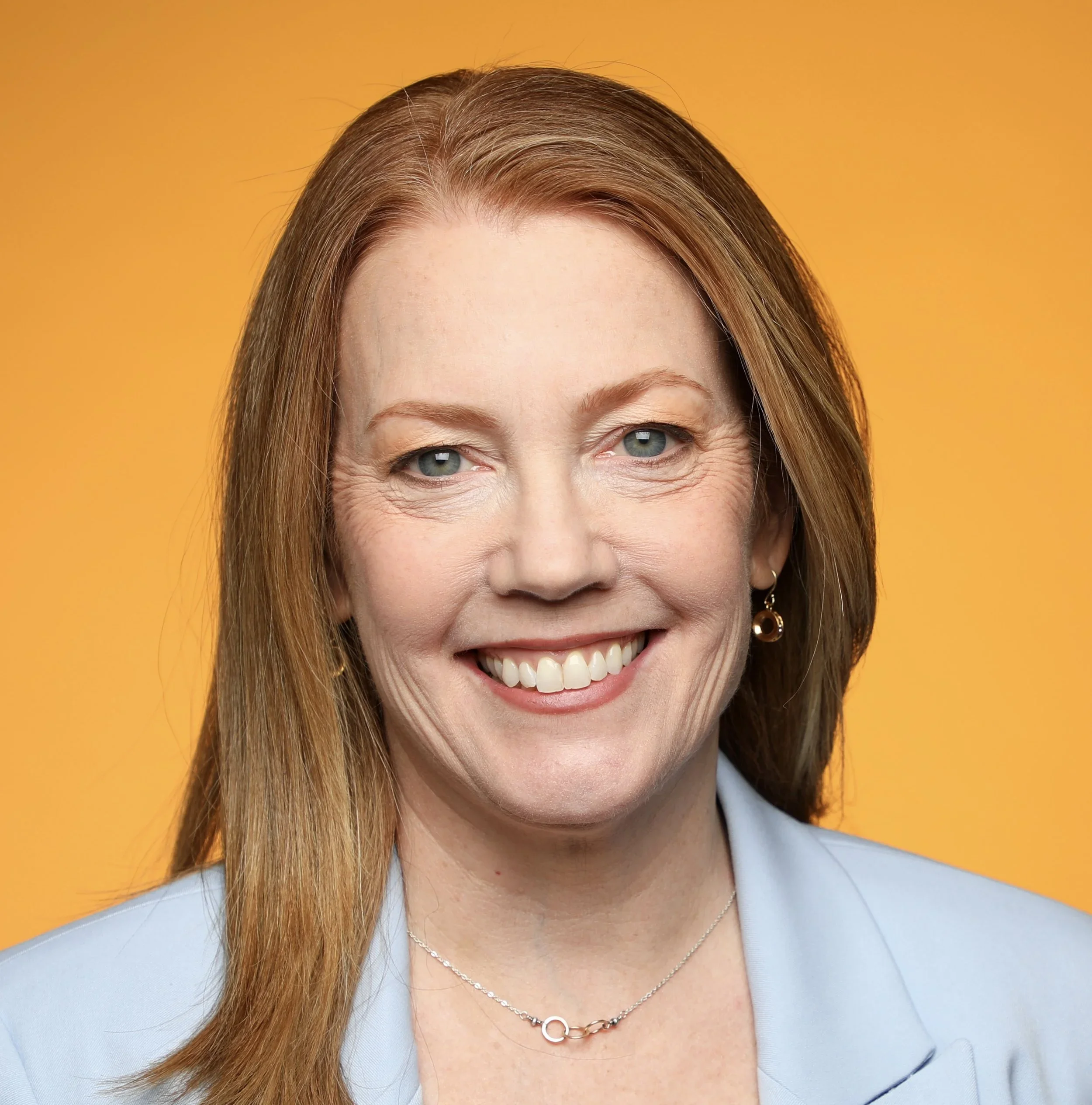 Expert EOS implementer Beth Fahey with red hair and blue eyes smiling, wearing a light blue blazer, earrings, and a necklace, against an orange background.