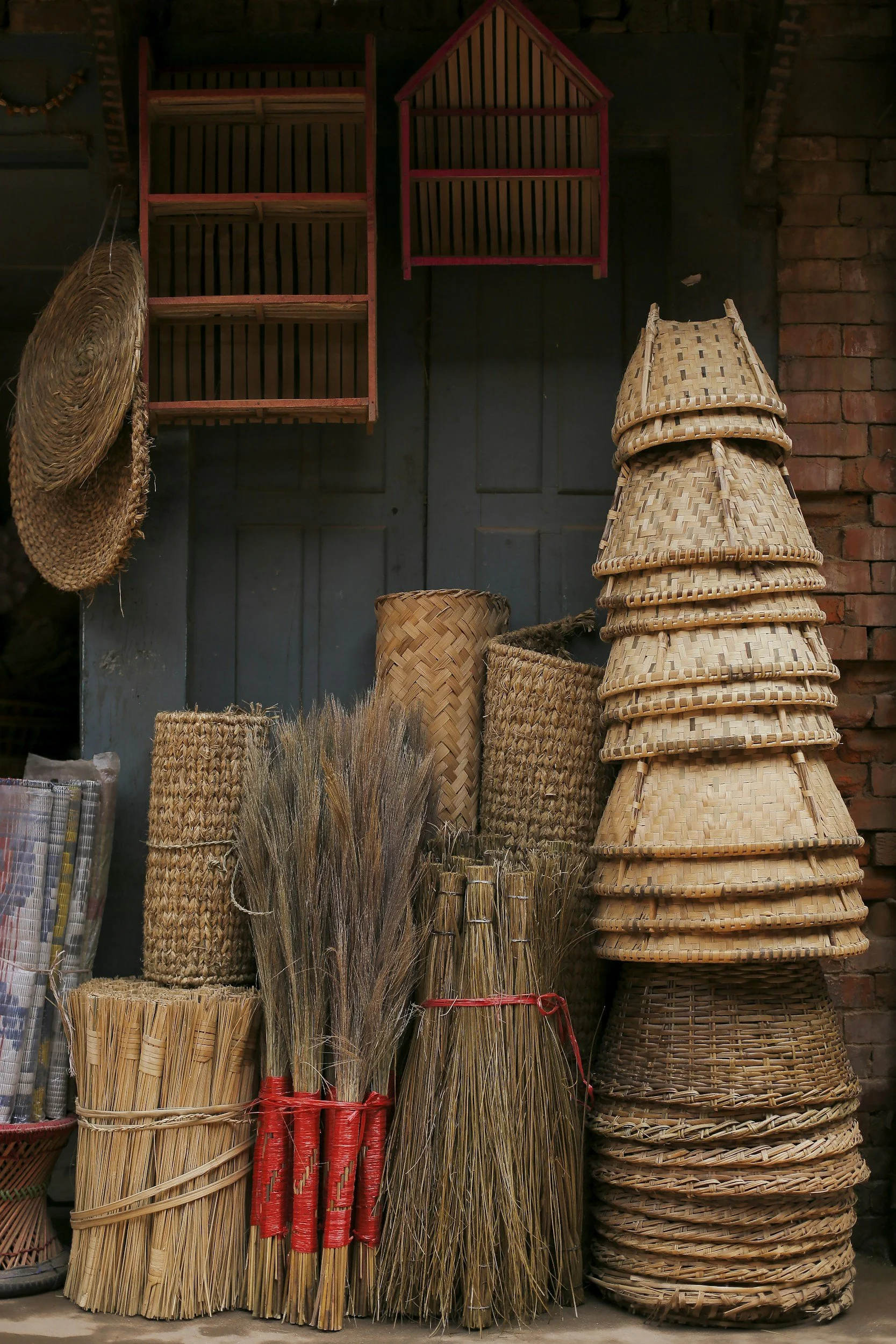 Stacks of woven baskets and brooms made from natural materials, displayed against a dark wall, with some hanging on the side and others piled in front.